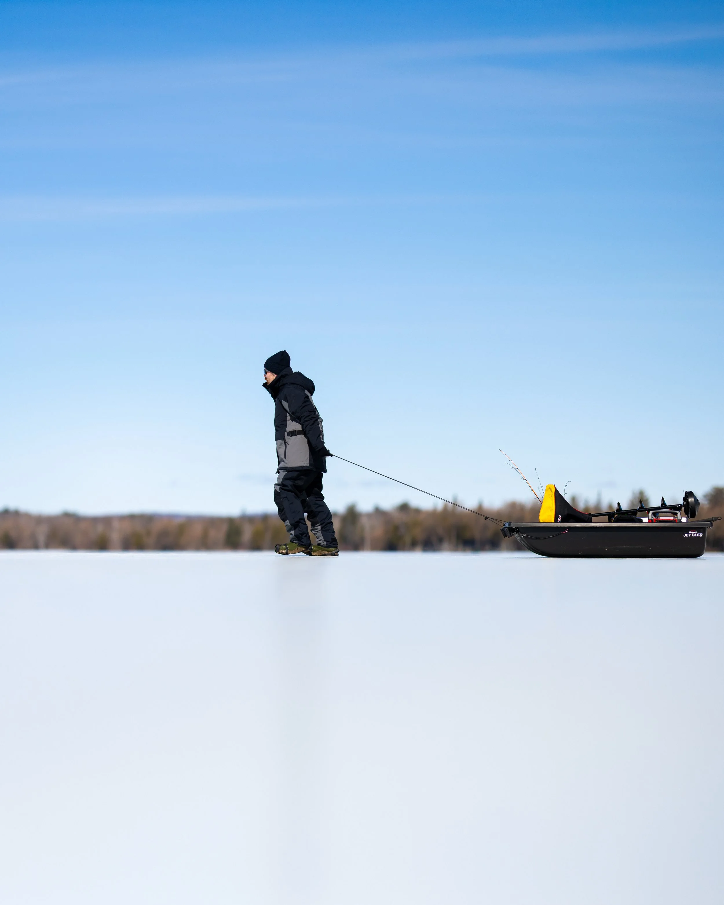 Person in winter clothing pulling a sled with fishing gear across frozen snow-covered landscape under blue sky.