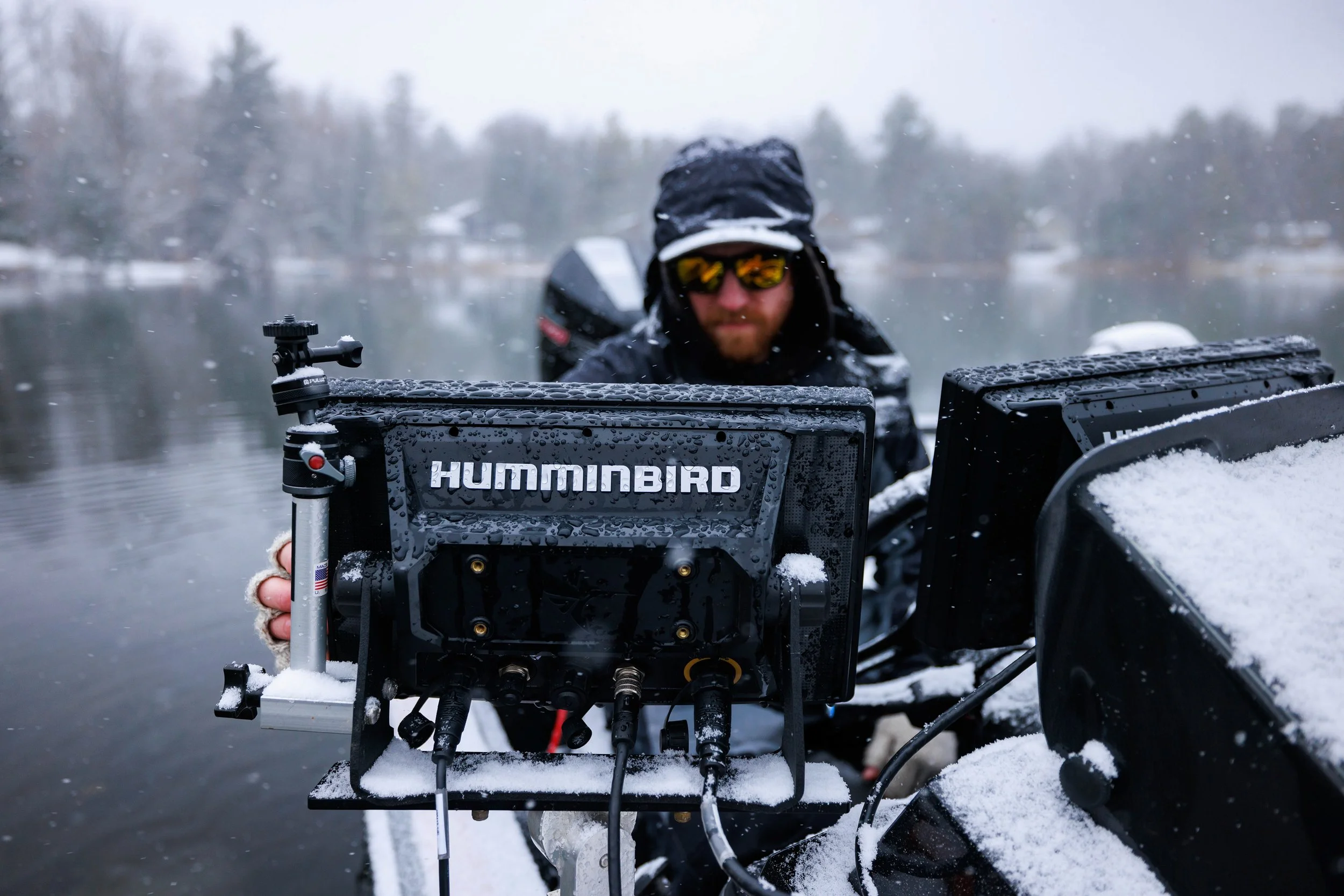 Man on a boat with Humminbird fish finder in a snowy outdoor setting, water and trees in the background.