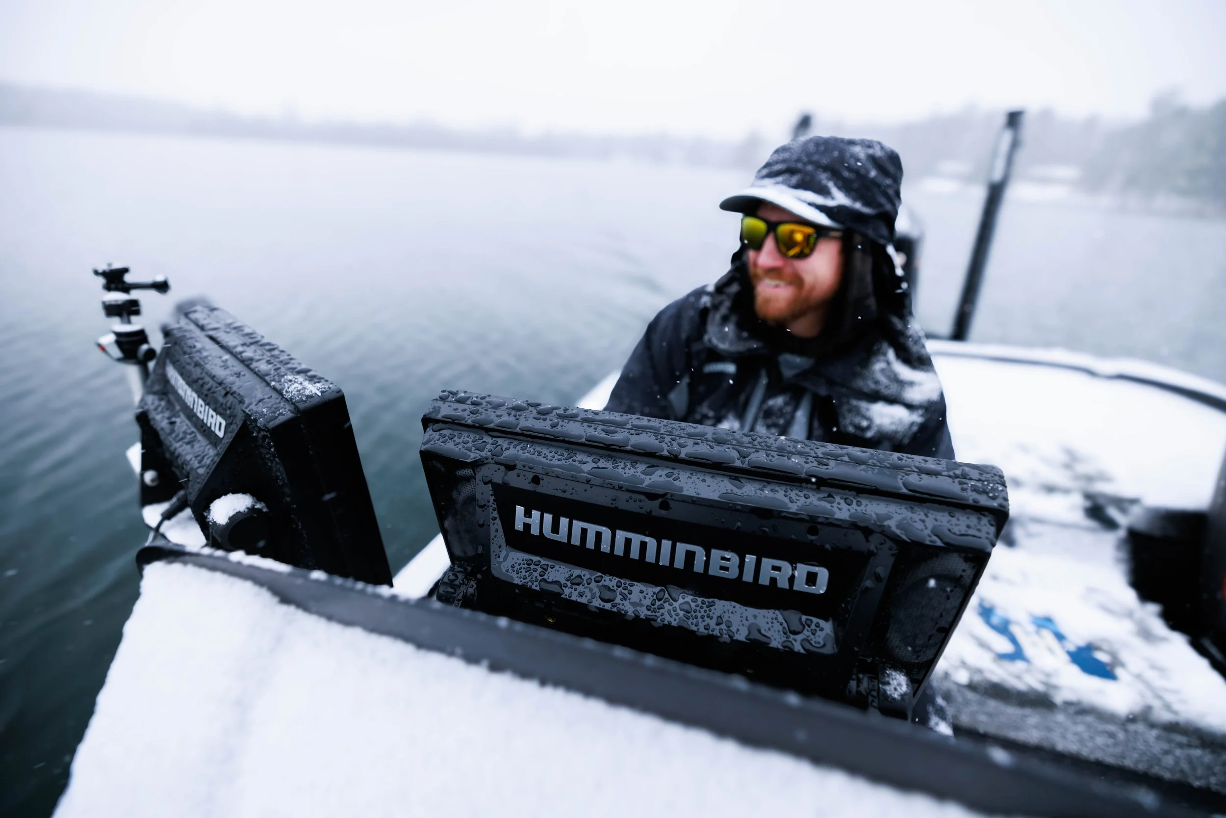 A man wearing sunglasses, a black hat, and a winter jacket is on a boat in a snowy, icy lake, smiling, with two Humminbird fish finders in front of him.