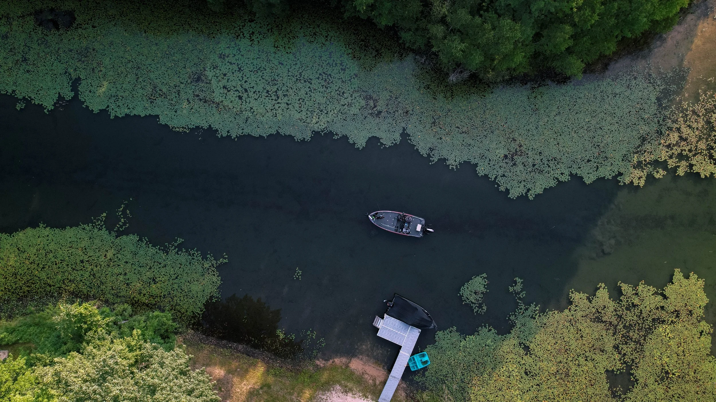 An aerial view of a small body of water surrounded by lush green trees and vegetation, with a dock extending into the water. A boat is anchored near the dock, and a smaller boat is tied to the dock. The water is dark and calm, with patches of floatin