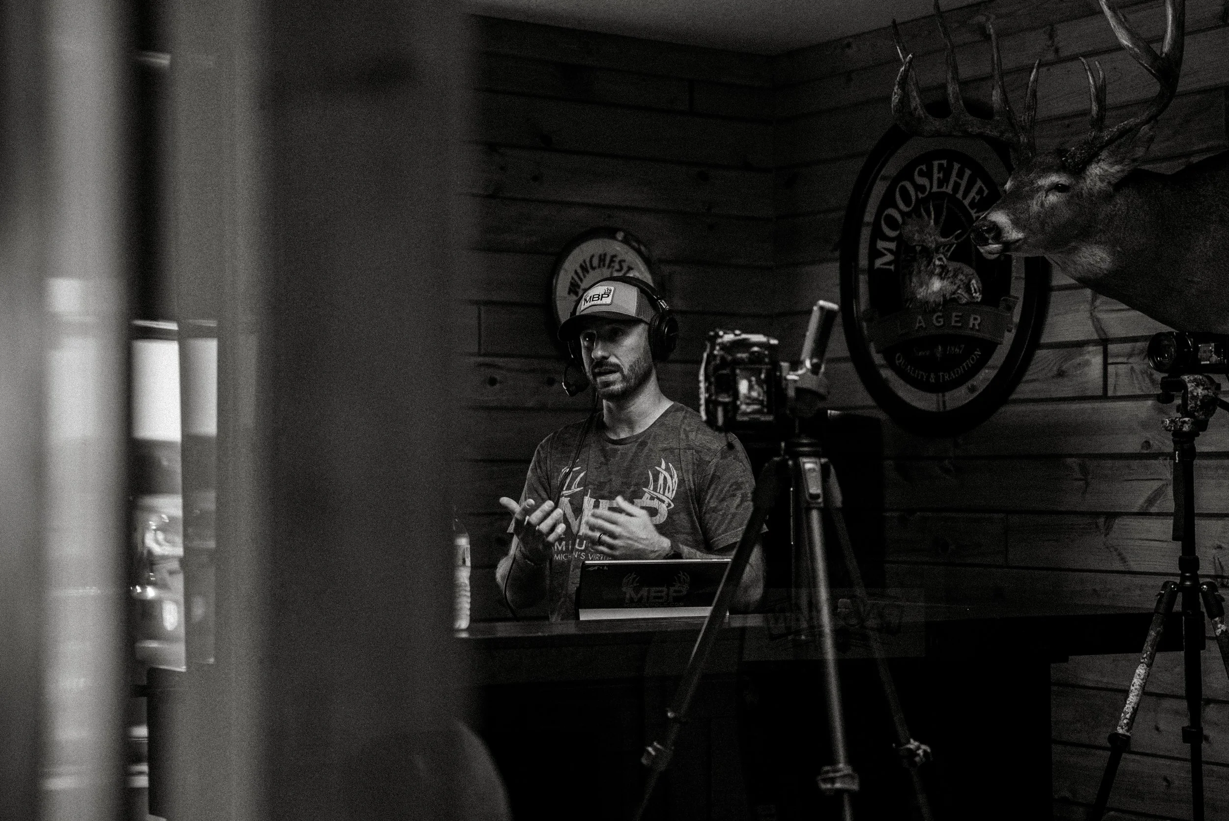 A man wearing a cap and headphones speaking into a microphone in a rustic wood-paneled room, with a mounted deer head and a Moosehead Beverage logo on the wall behind him.