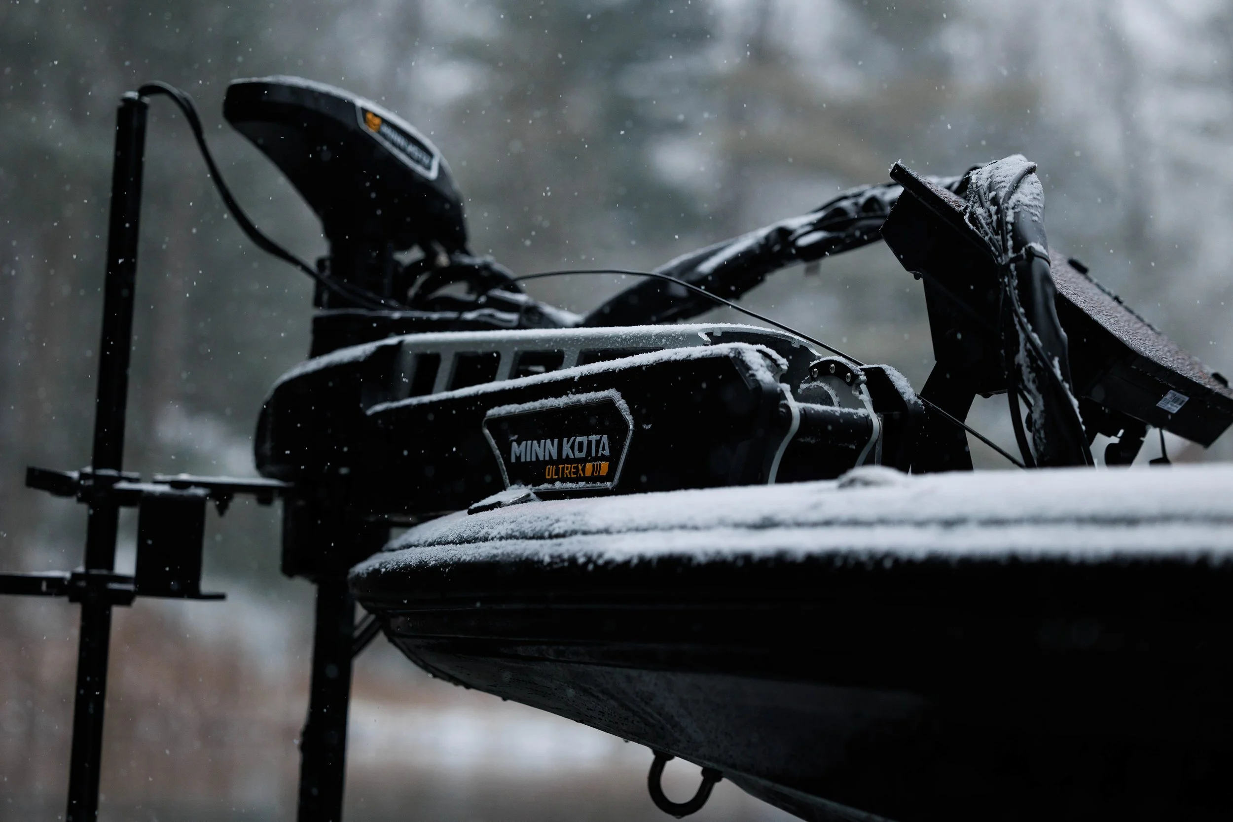 A snow-covered boat with a Minn Kota Ultrex trolling motor resting on a snow-covered dock, with a blurred natural background.