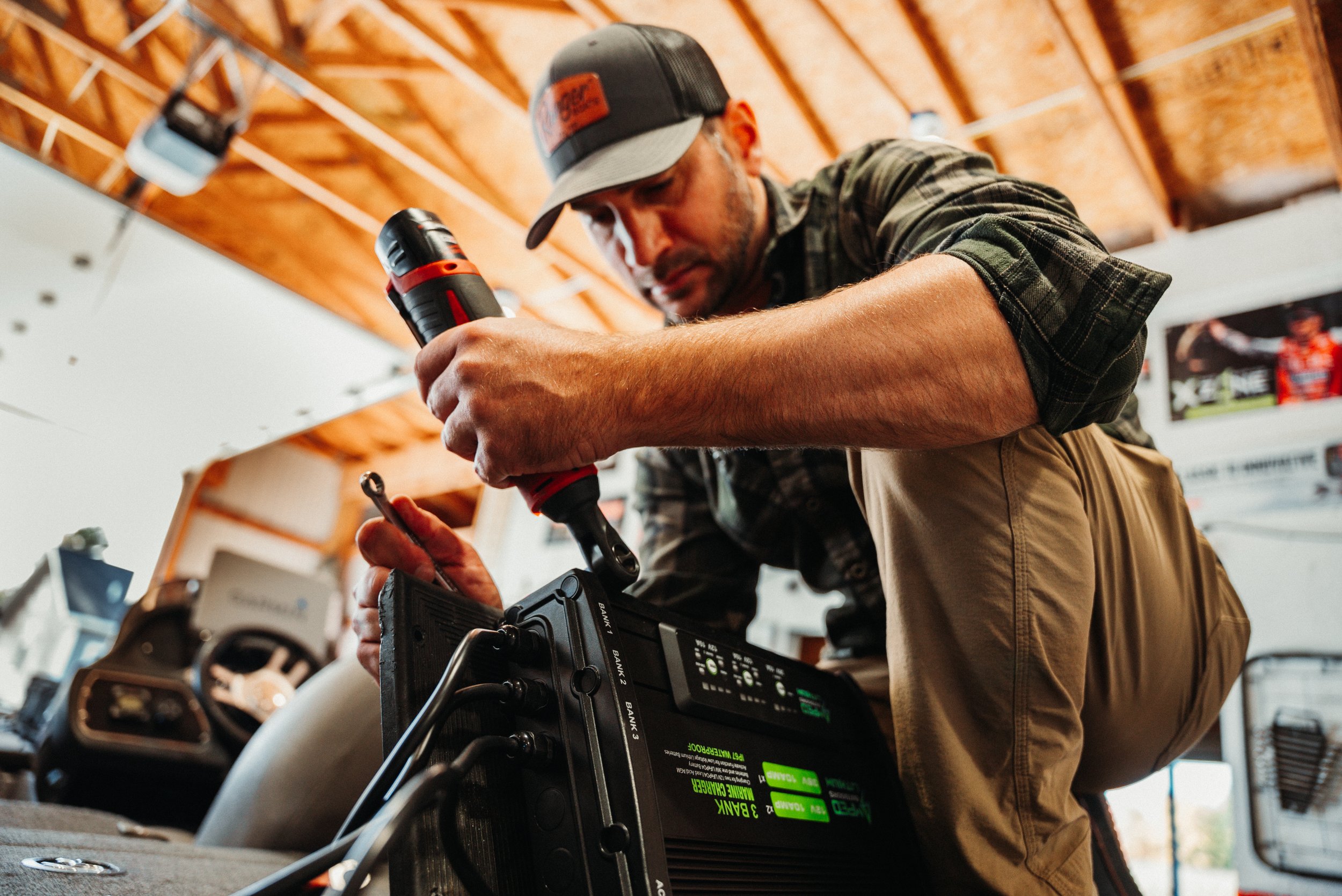A man kneeling on the floor, using a cordless drill to repair electronic equipment inside a garage or workshop. He is wearing a plaid shirt, khaki pants, and a baseball cap.