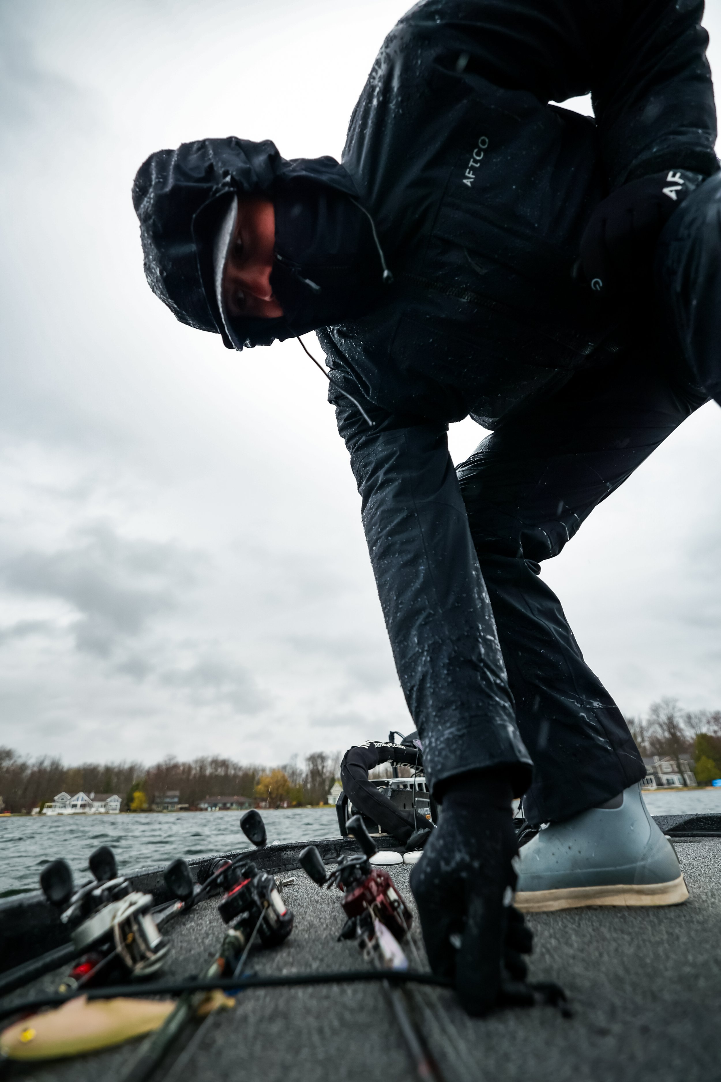 Person dressed in waterproof fishing gear on a boat, preparing fishing rods near a body of water on a cloudy day.