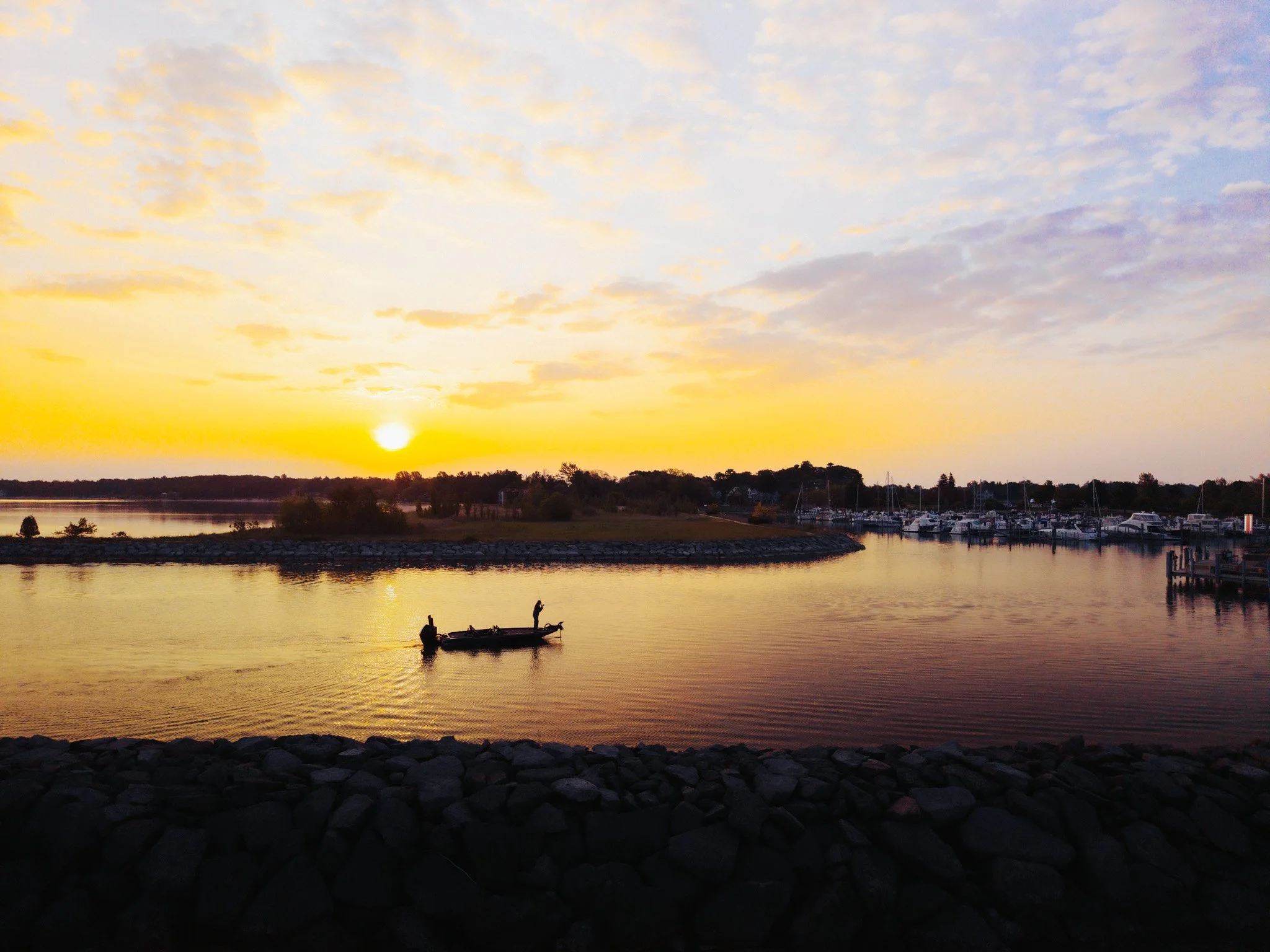 A person fishing from a small boat in calm water during sunset, with a marina and a tree-lined shoreline in the background.