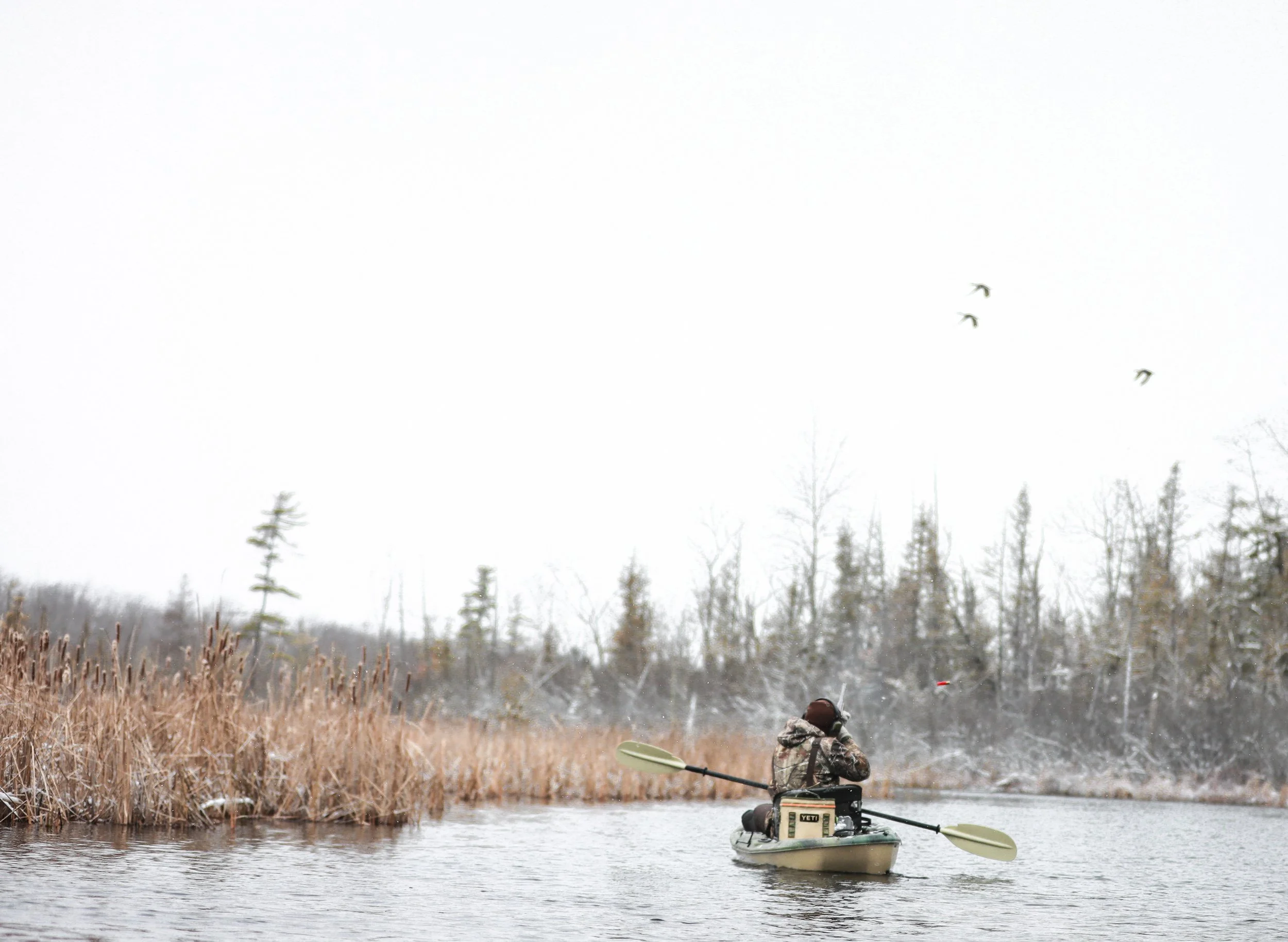 Person in camouflage clothing kayaking on a lake during winter with bare trees and reeds along the shore, and birds flying in the sky.