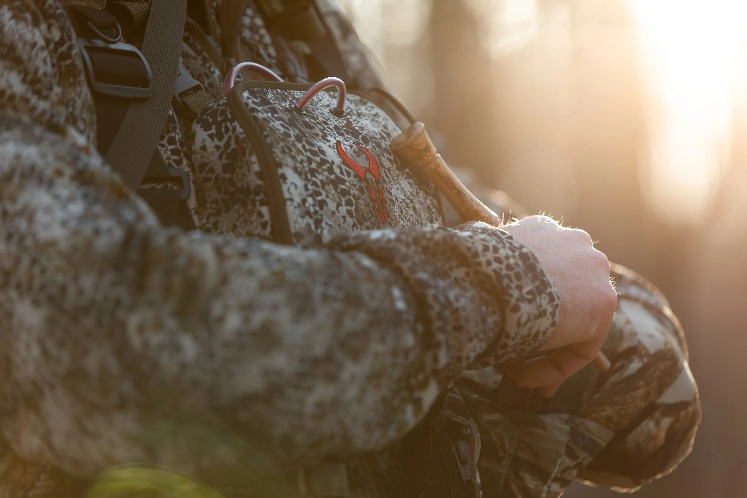 Close-up of a person wearing a camouflage jacket holding a hunting rifle near a backpack with a red deer antler logo during sunrise or sunset.