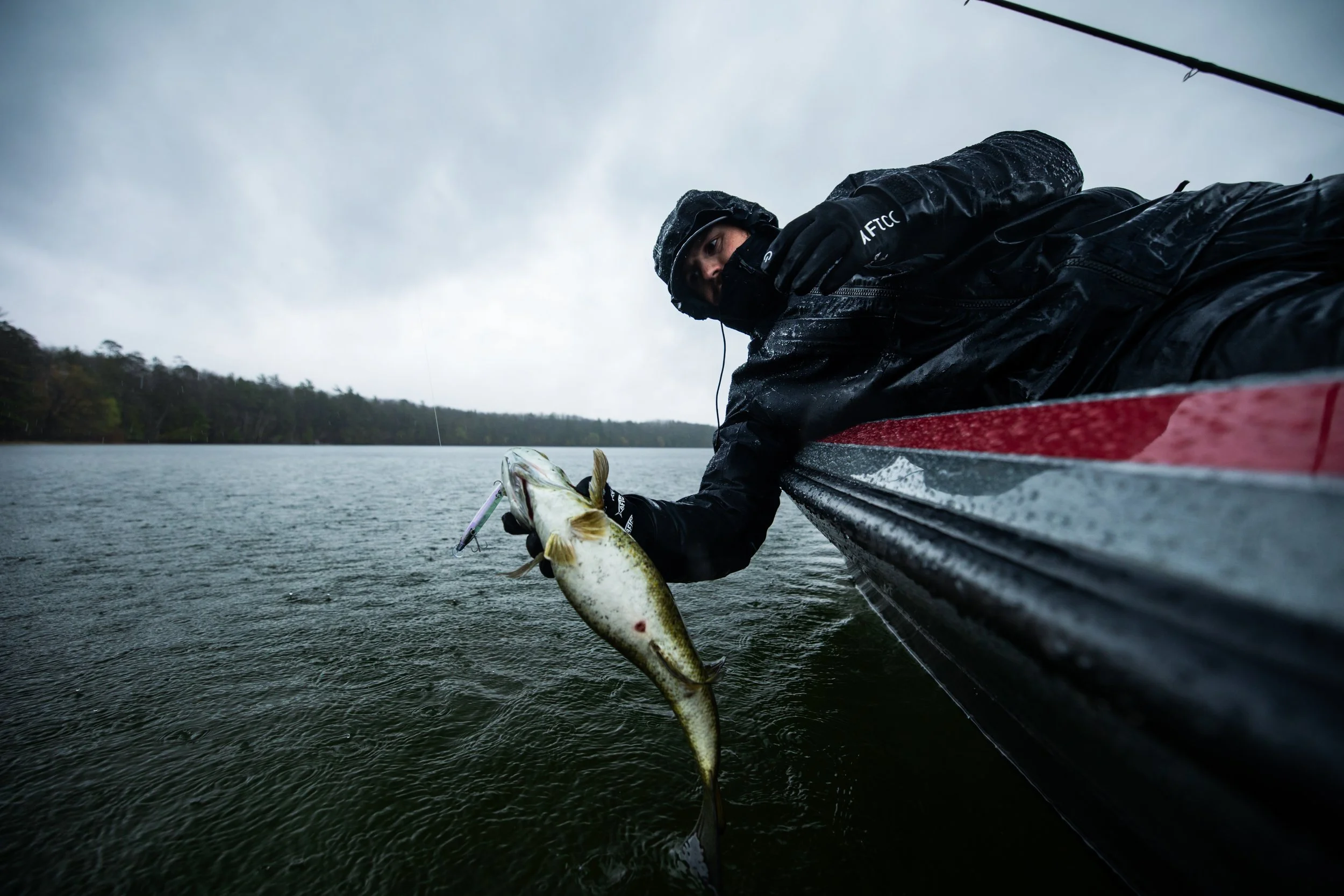 Person in waterproof gear lying on a boat, holding a large fish caught from the water with a fishing lure in its mouth, during overcast weather.
