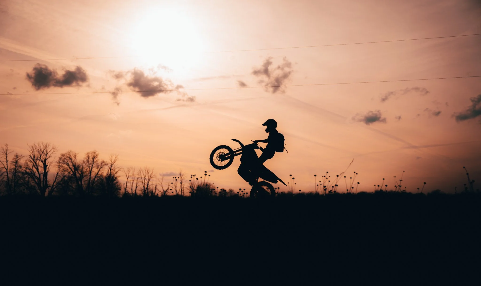 Silhouette of a person performing a wheelie on a dirt bike at sunset with trees and clouds in the background.