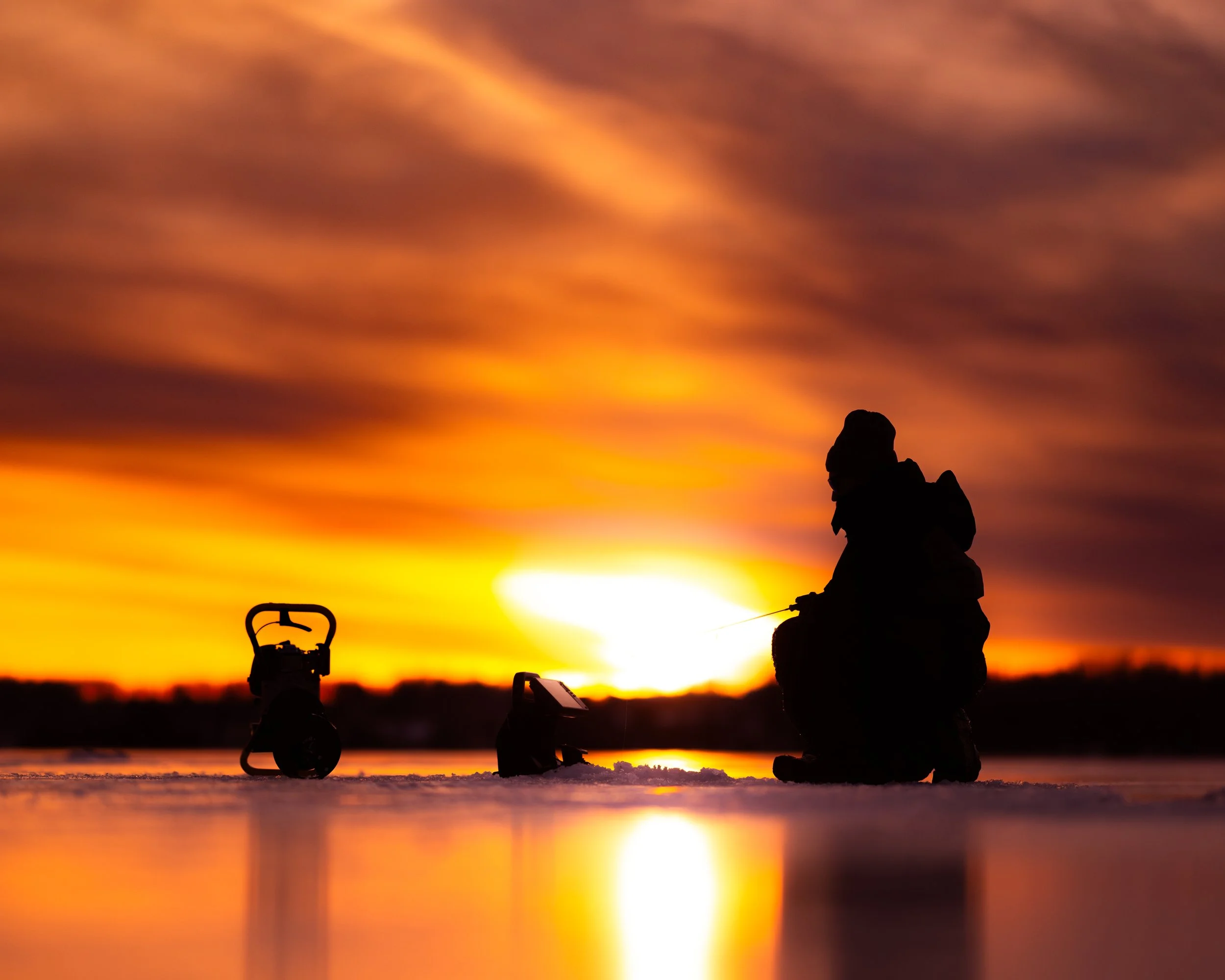 Silhouette of a person fishing at sunset with fishing gear on a frozen surface.