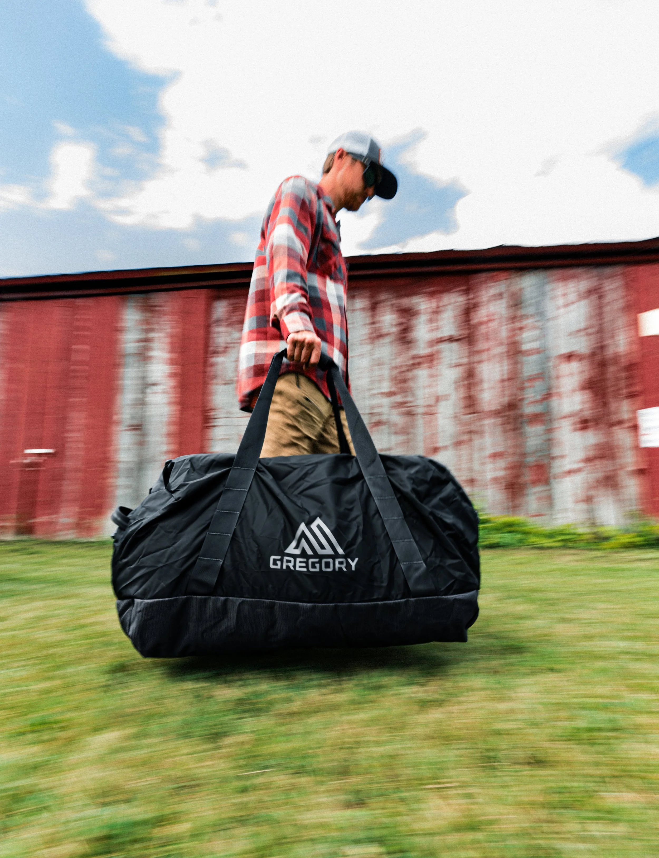 A man in a plaid shirt, beige pants, sunglasses, and a baseball cap walking on grass carrying a large black duffel bag with the logo 'Gregory' on it, with a red barn and cloudy sky in the background.
