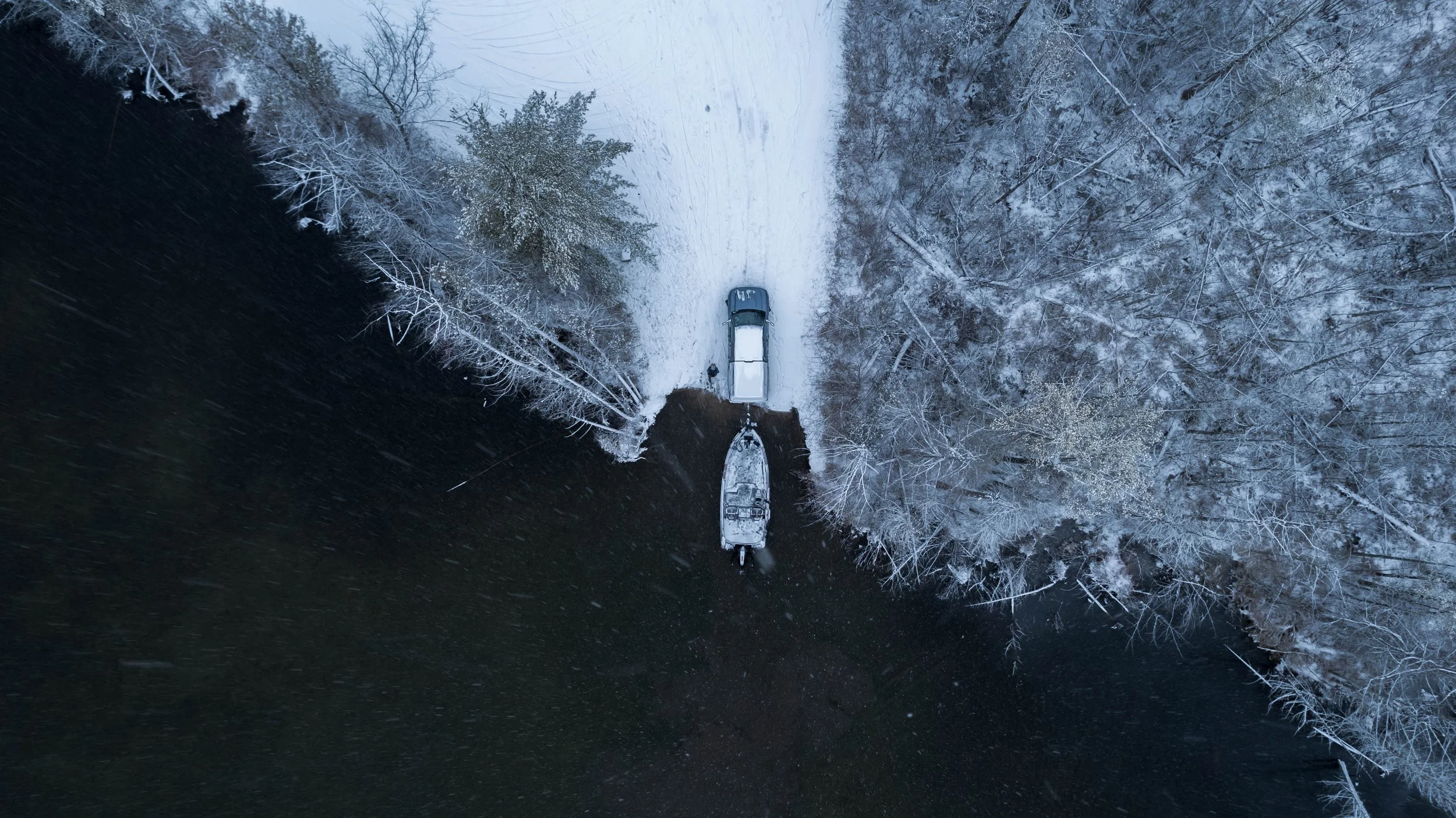 Aerial view of a snow-covered area with a car parked next to a boat on a trailer, surrounded by snow-laden trees.