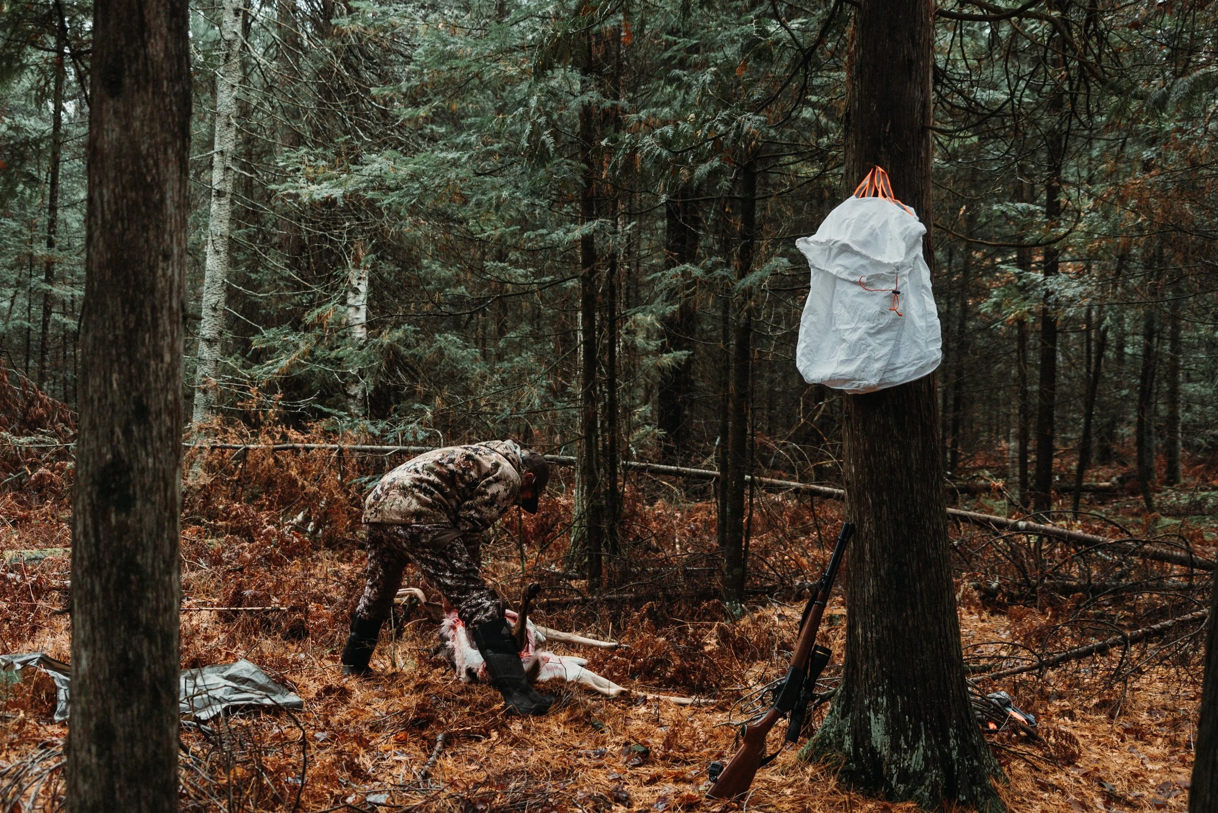 A person wearing camouflage clothing in a forest, preparing a hunted animal on the ground. A rifle leans against a tree with a white bag hanging from a branch nearby.