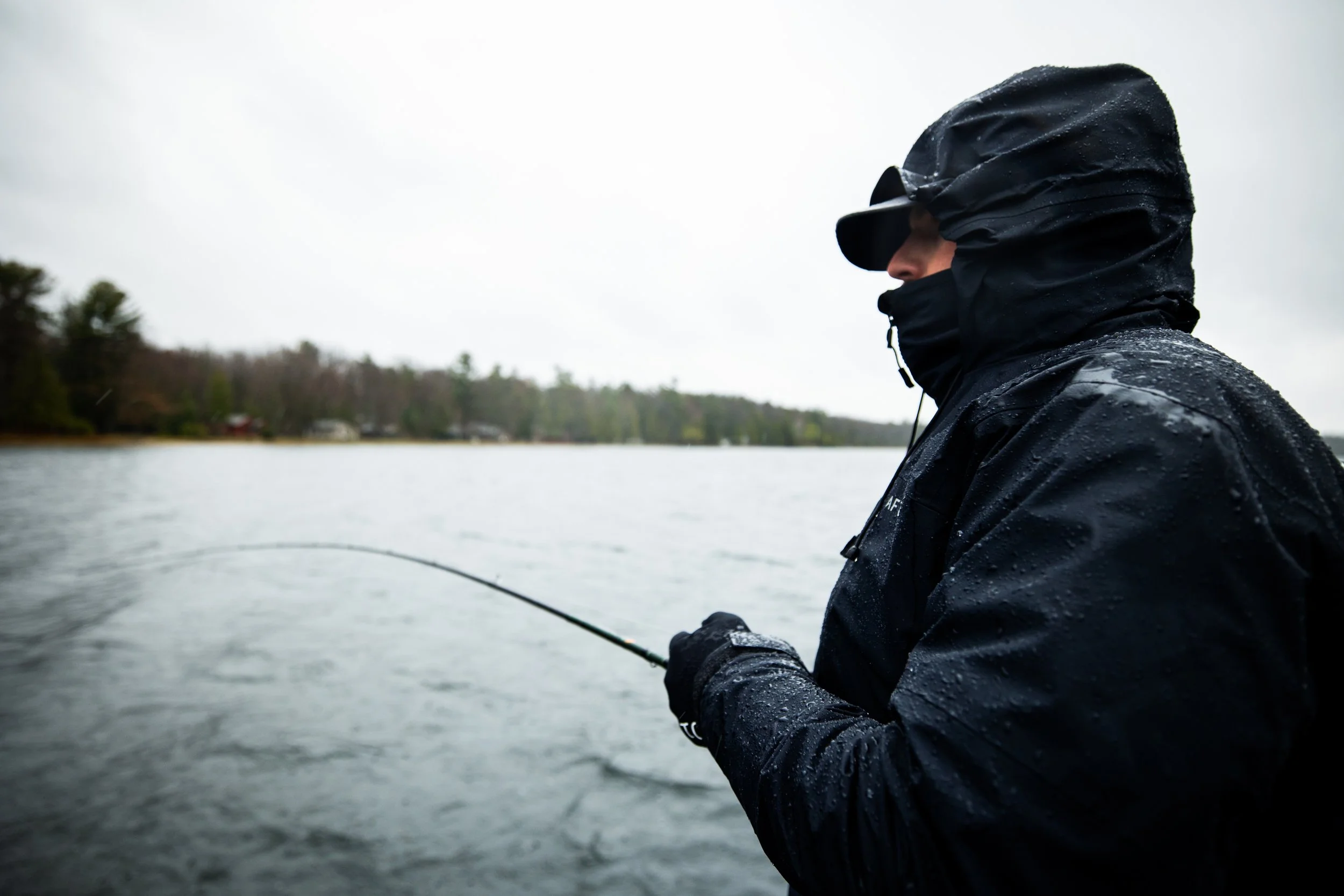Person in black rain jacket and hat fishing in a lake during overcast weather.