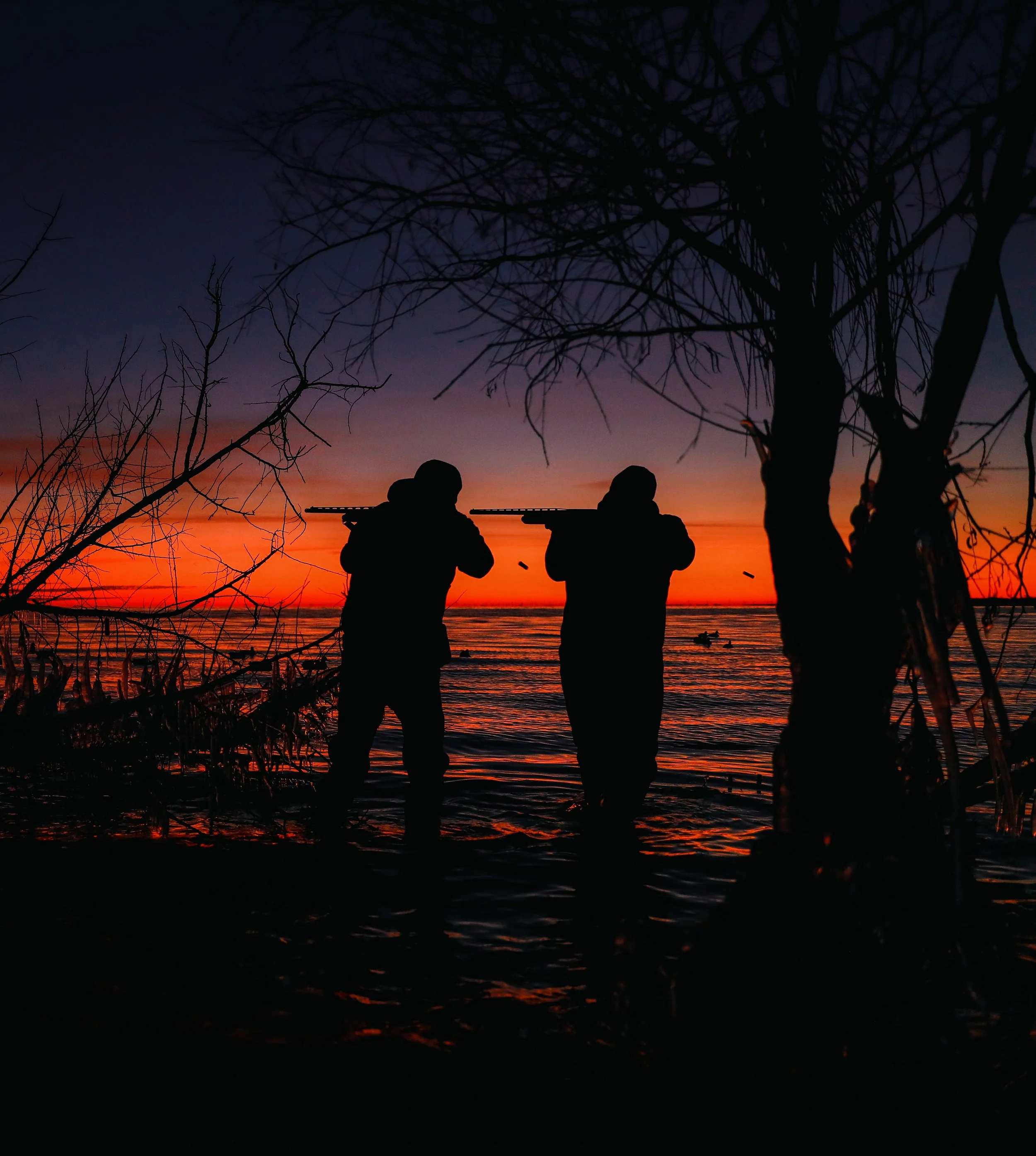Silhouettes of two people holding rifles standing by a river at sunset with trees in the foreground.
