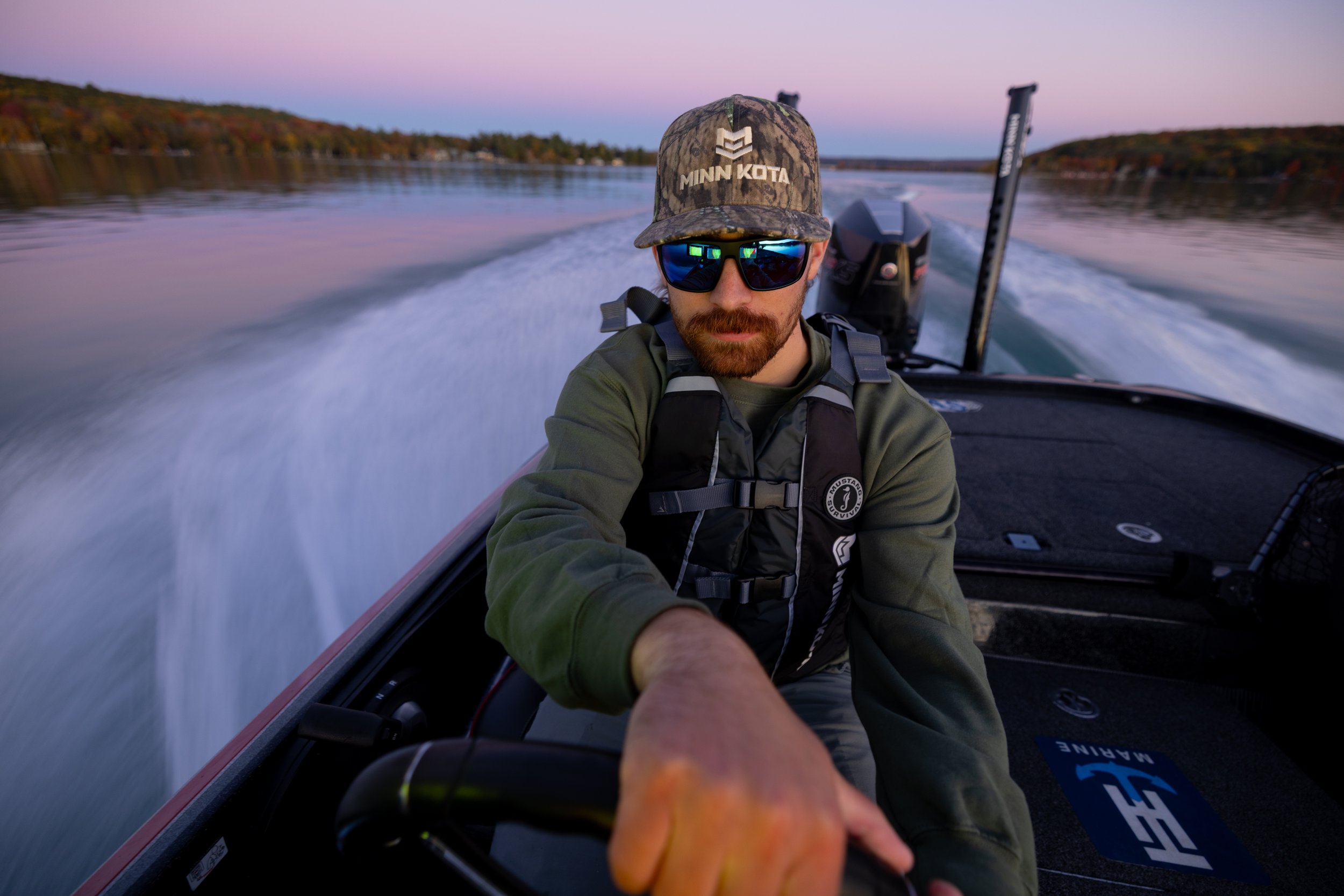 A man with a beard, wearing sunglasses, a Minnesota cap, and a green jacket, is taking a selfie while riding a speedboat on a lake during sunset, with trees and hills in the background.