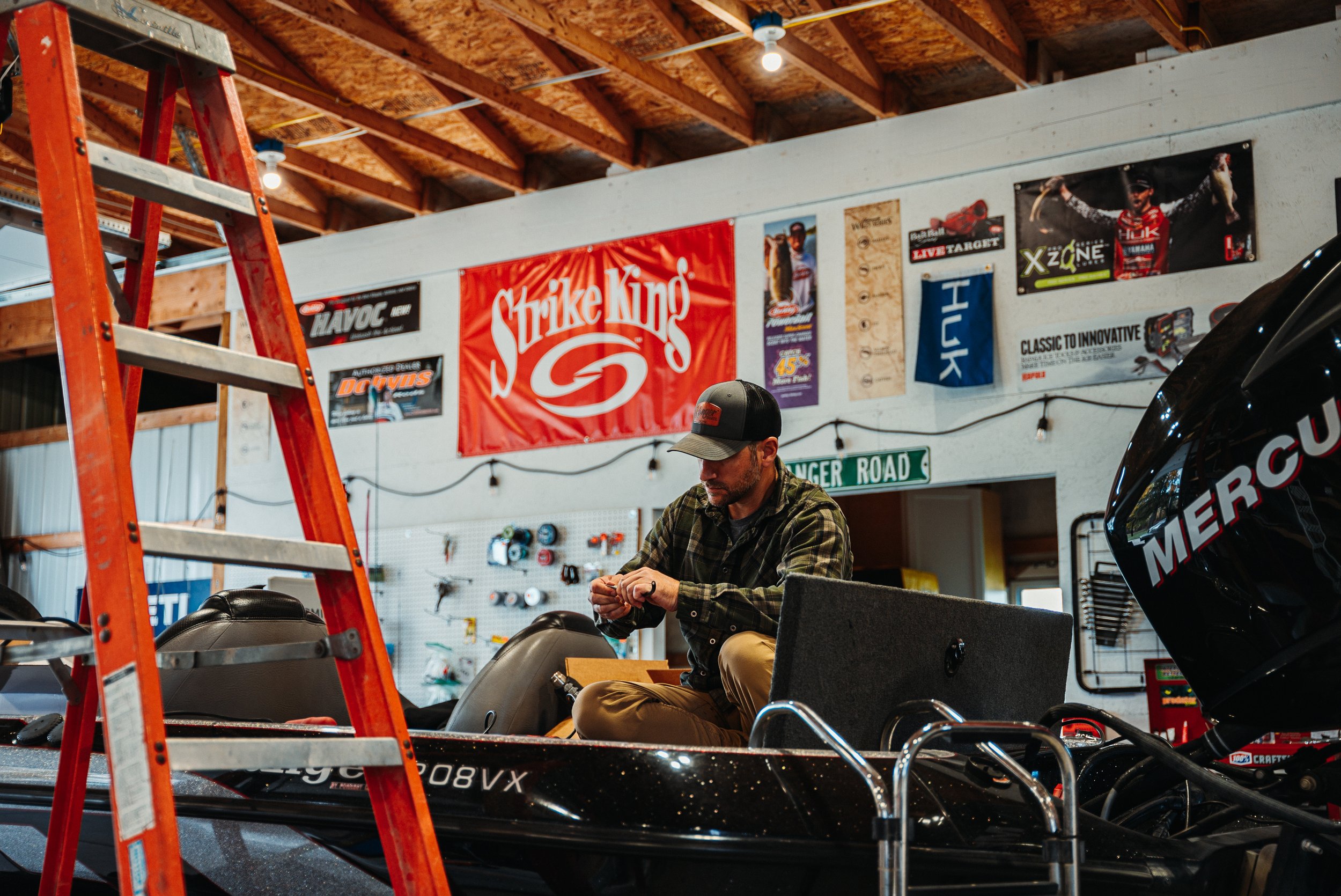A man sitting in a workshop or garage next to a boat with a Mercury outboard motor. The man is wearing a cap and a plaid shirt, working with his hands. The background has various posters and banners, including a large red Strike King banner, and a si
