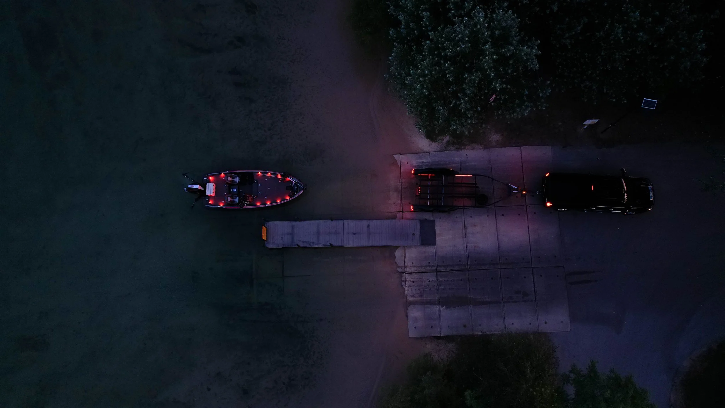 An aerial view of a boat with red lights on a trailer attached to a black vehicle parked on a concrete area near trees at night.