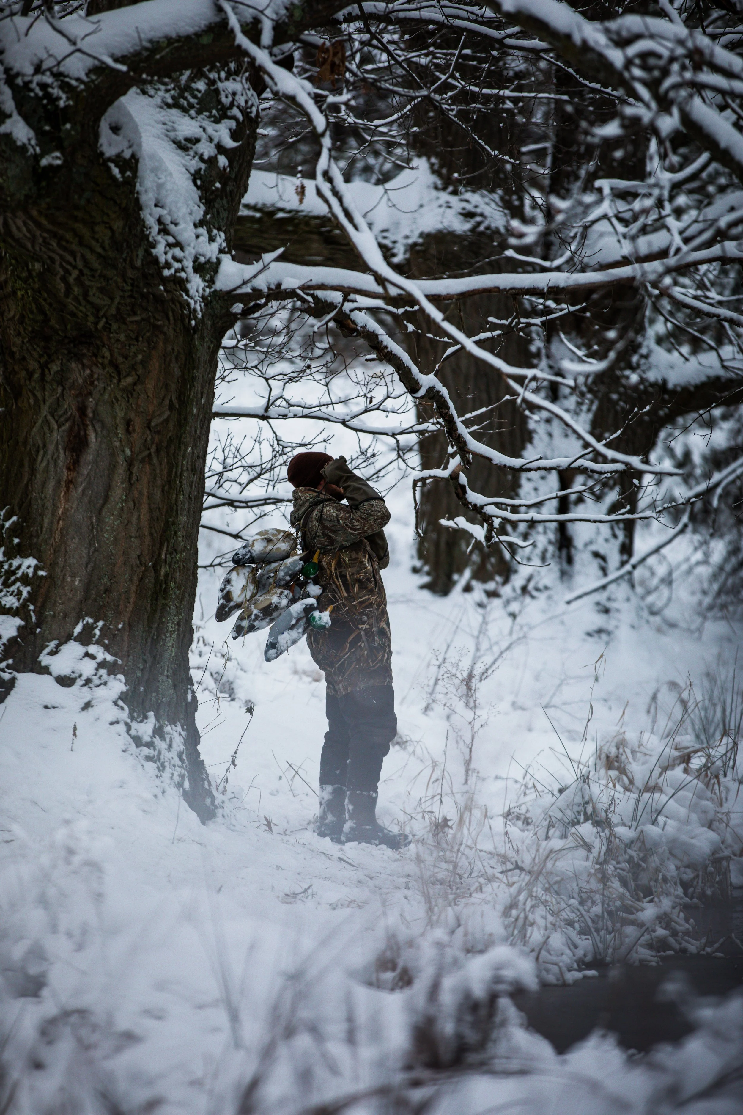 Person dressed in camouflage clothing standing near a snow-covered tree with a backpack filled with hunting boots in a winter landscape.