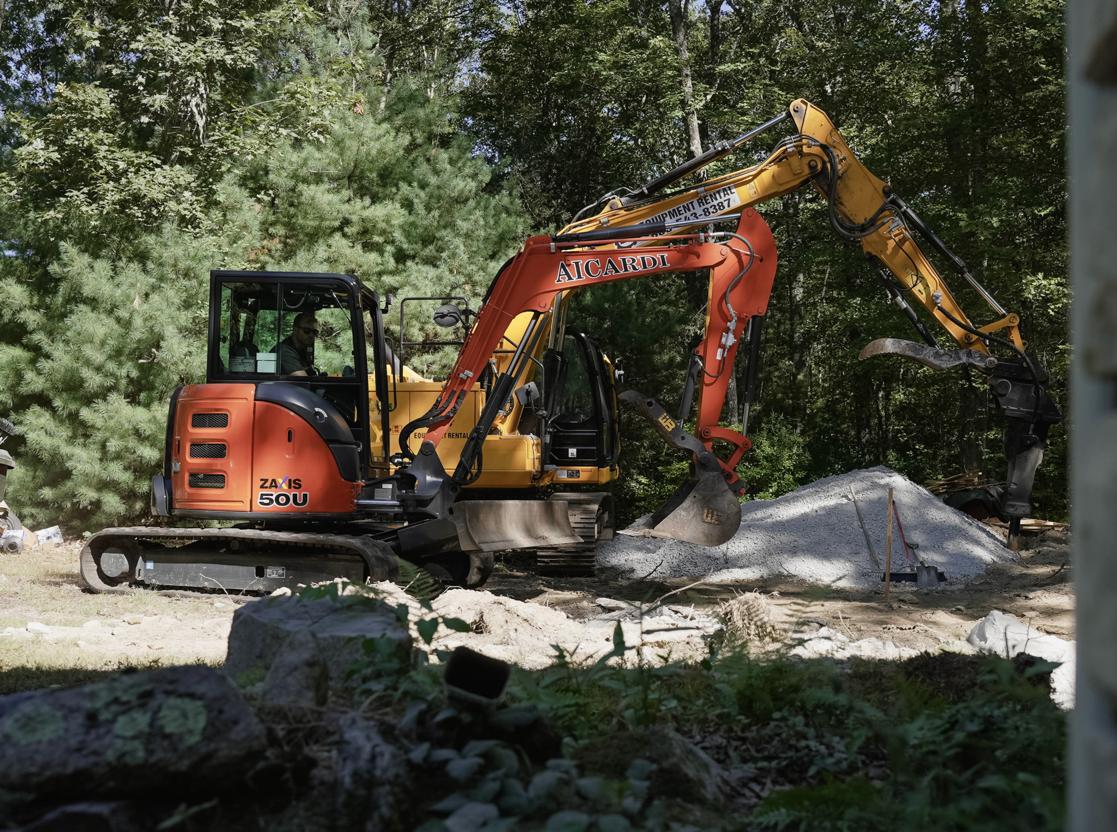 A construction excavator digging in a forested area, with trees in the background and a pile of dirt in front.