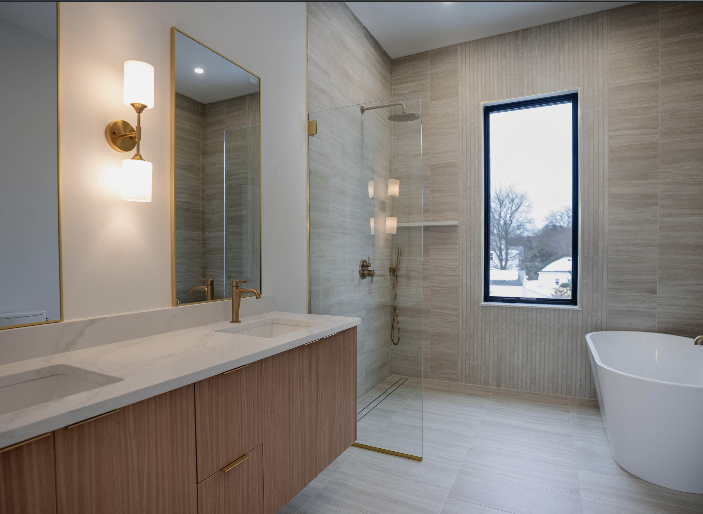 Modern bathroom with double vanity, large mirror, wall sconce, walk-in shower with glass door, window showing snowy outdoor scene, and freestanding bathtub.