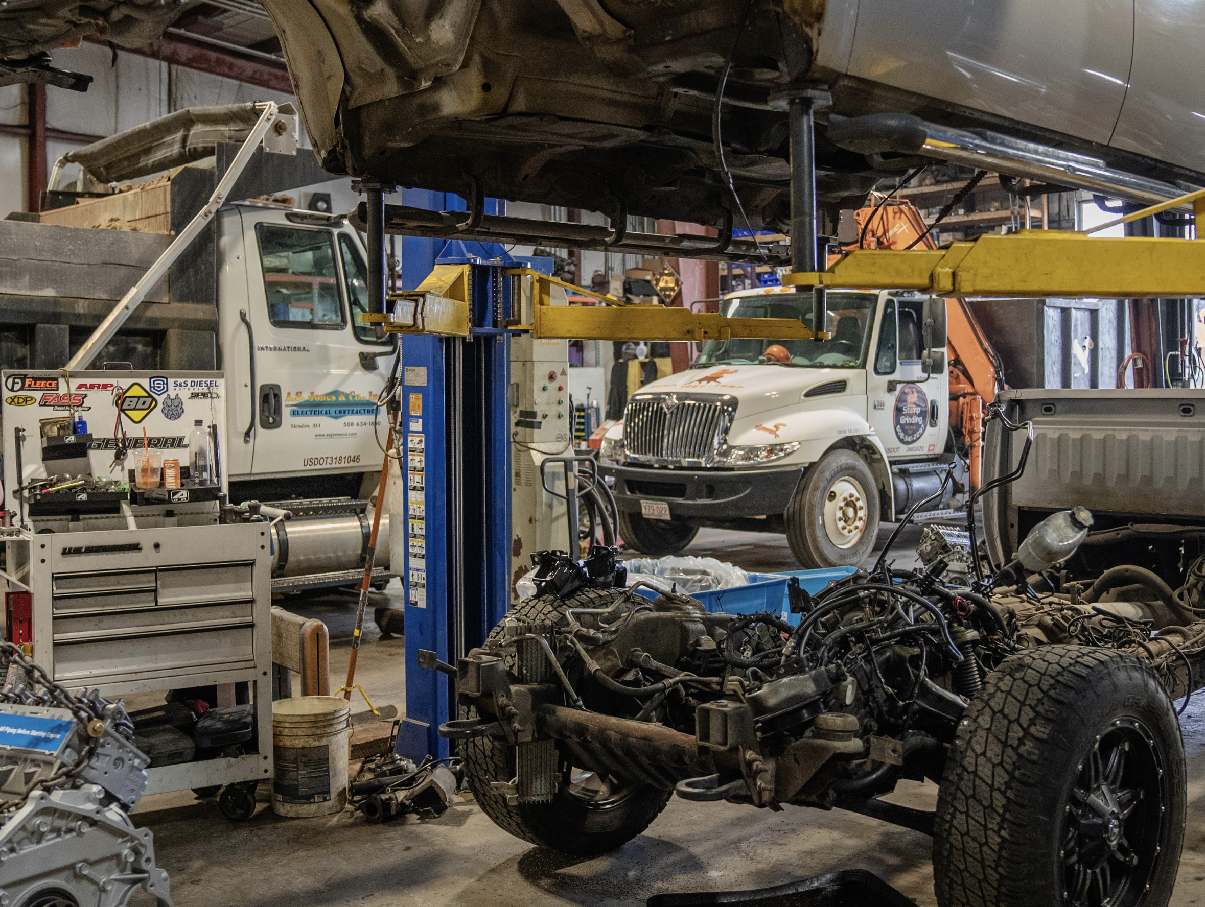 Auto repair shop with vehicles, tools, and equipment, including a partially disassembled car with visible engine and a white truck on a lift.