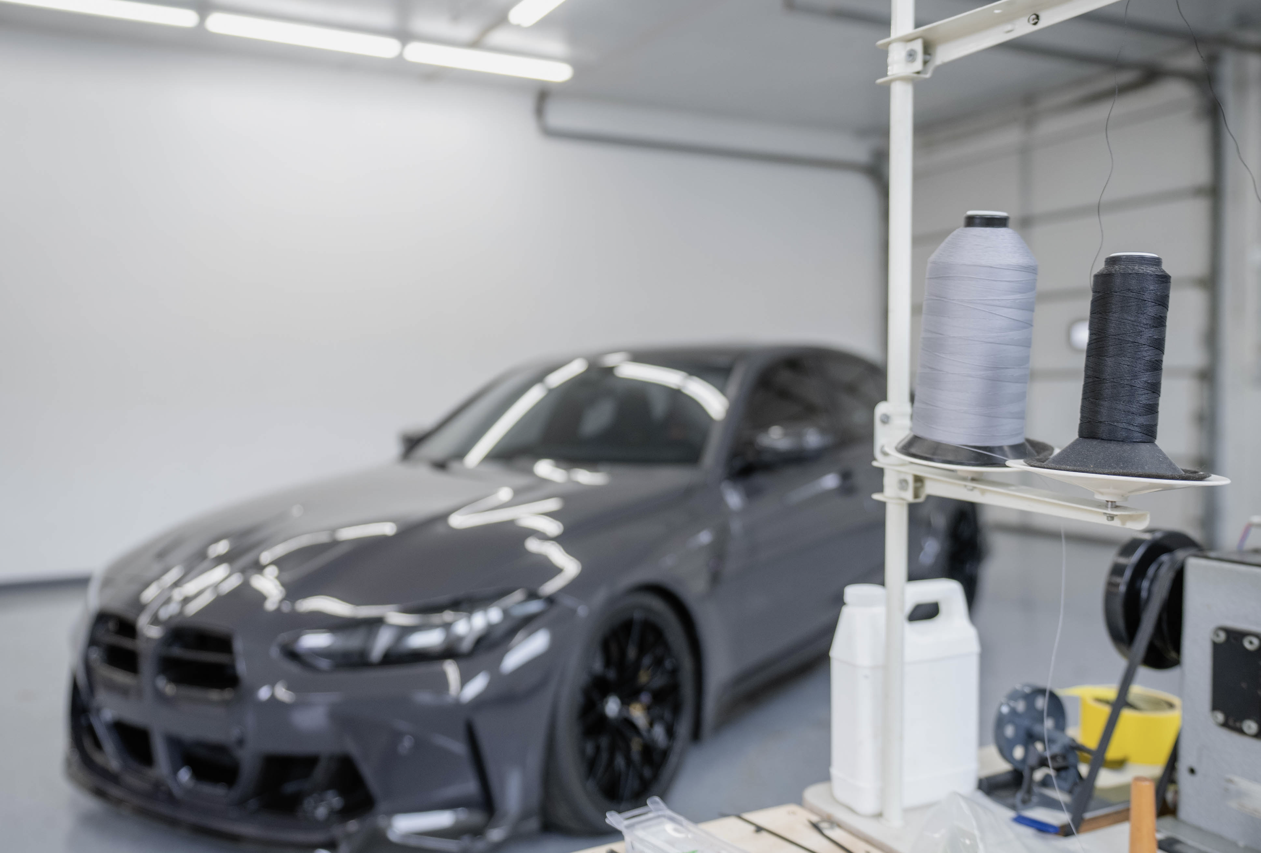 A sleek black sports car inside a workshop or garage with sewing materials, such as spools of thread, in the foreground.