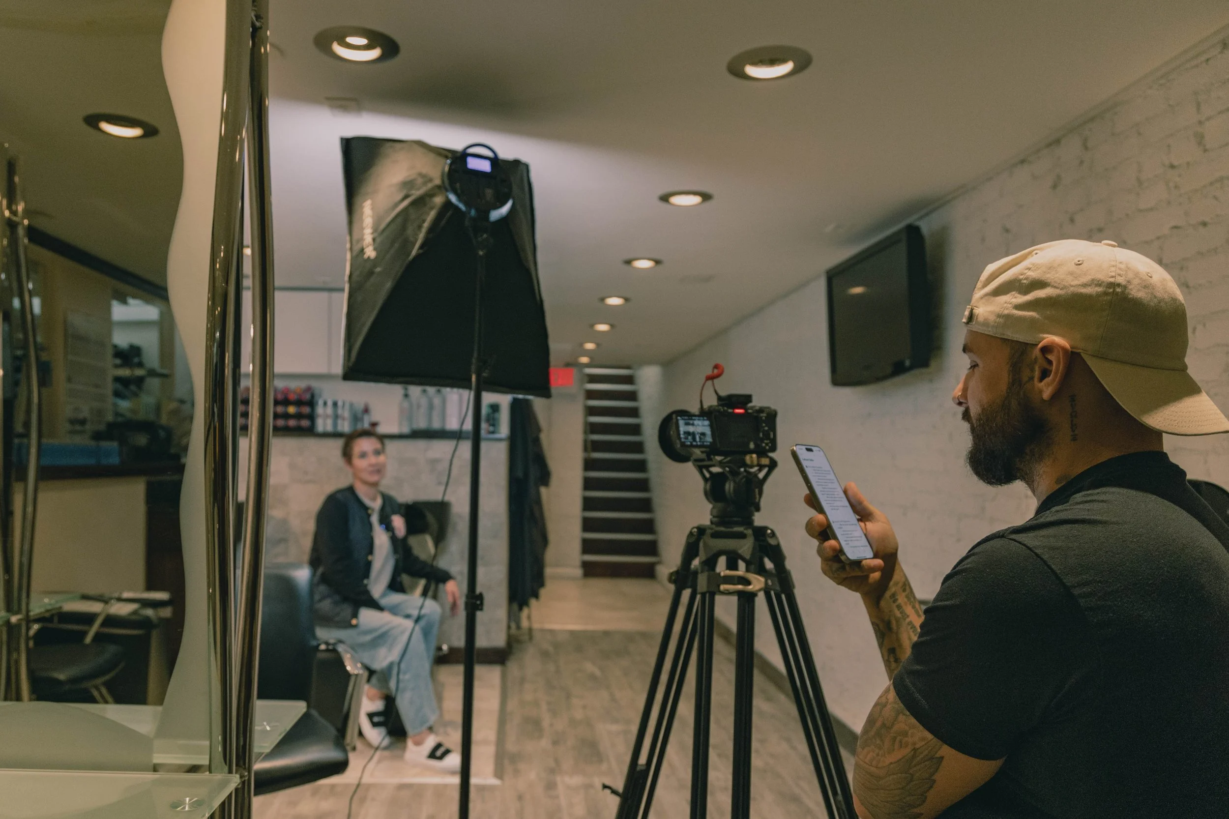 A man with tattoos wearing a beige cap and black shirt is filming a woman sitting on a chair in a well-lit, modern indoor space with white brick walls. The man is looking at his phone while managing a camera on a tripod, and there is a large studio light directed toward the woman for filming.