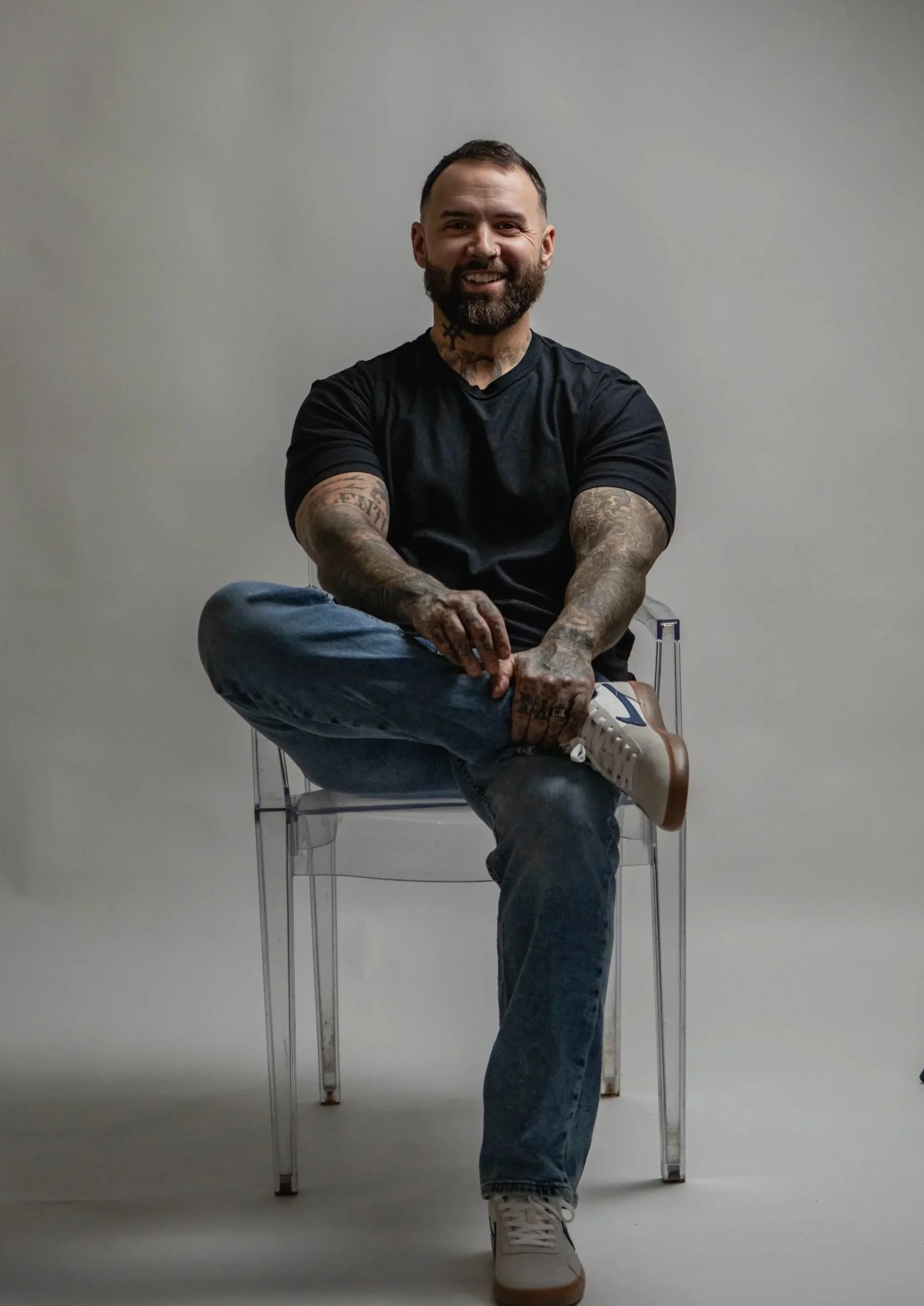 A tattooed man in a black t-shirt and blue jeans sitting cross-legged on a transparent chair against a plain light gray background, smiling at the camera.