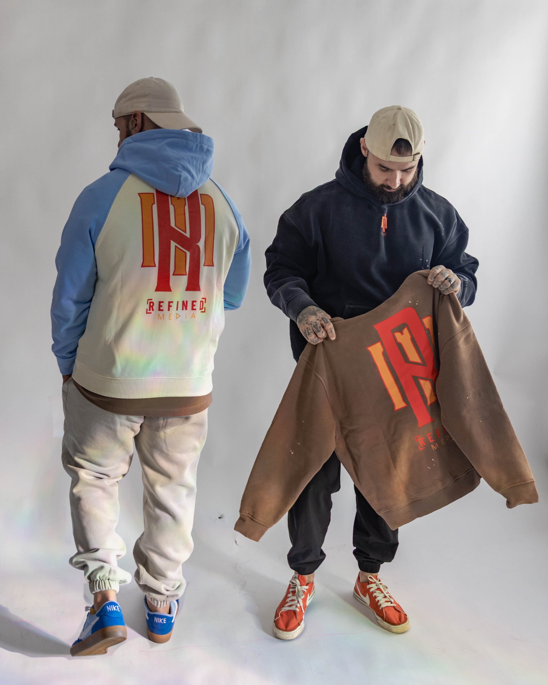 Two men standing against a plain white background. The man on the left faces away, wearing a beige cap, a light blue and cream hoodie with red and orange lettering and logo on the back, cream colored sweatpants, and blue Nike sneakers. The man on the right faces downward, holding a brown hoodie with similar red and orange lettering and logo, wearing a beige cap backwards, a black hoodie, black pants, and orange sneakers with white laces.