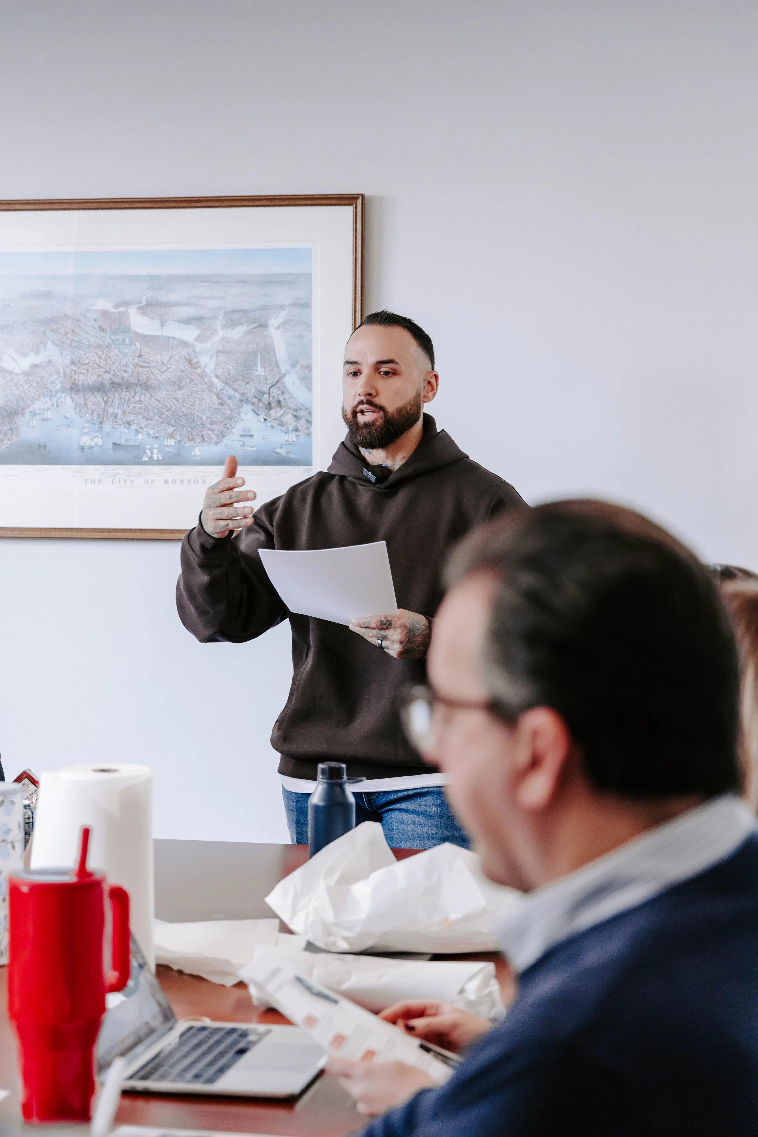 Man standing and speaking to a group of people in a meeting room, holding a paper, with a large framed map of Boston hanging on the wall behind him, and other attendees sitting at a table with laptops, papers, and drinks.