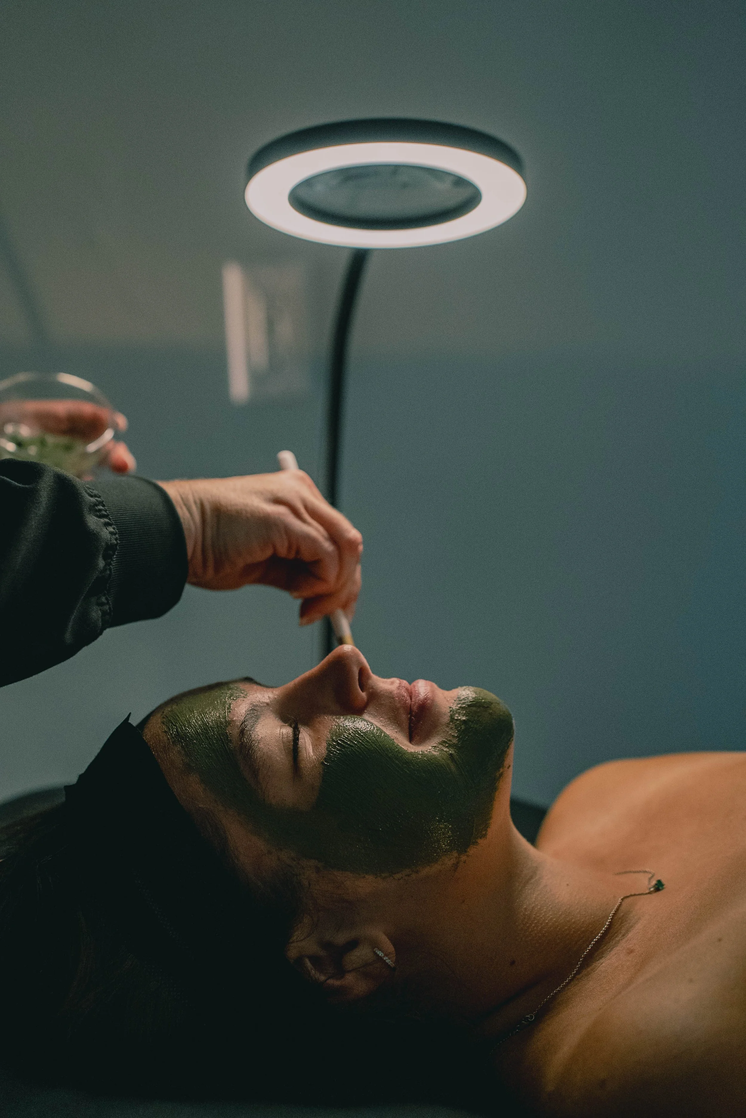 A woman receiving a facial treatment with a green mud mask, lying down with her eyes closed, in a spa or skincare clinic, under a modern overhead lamp.