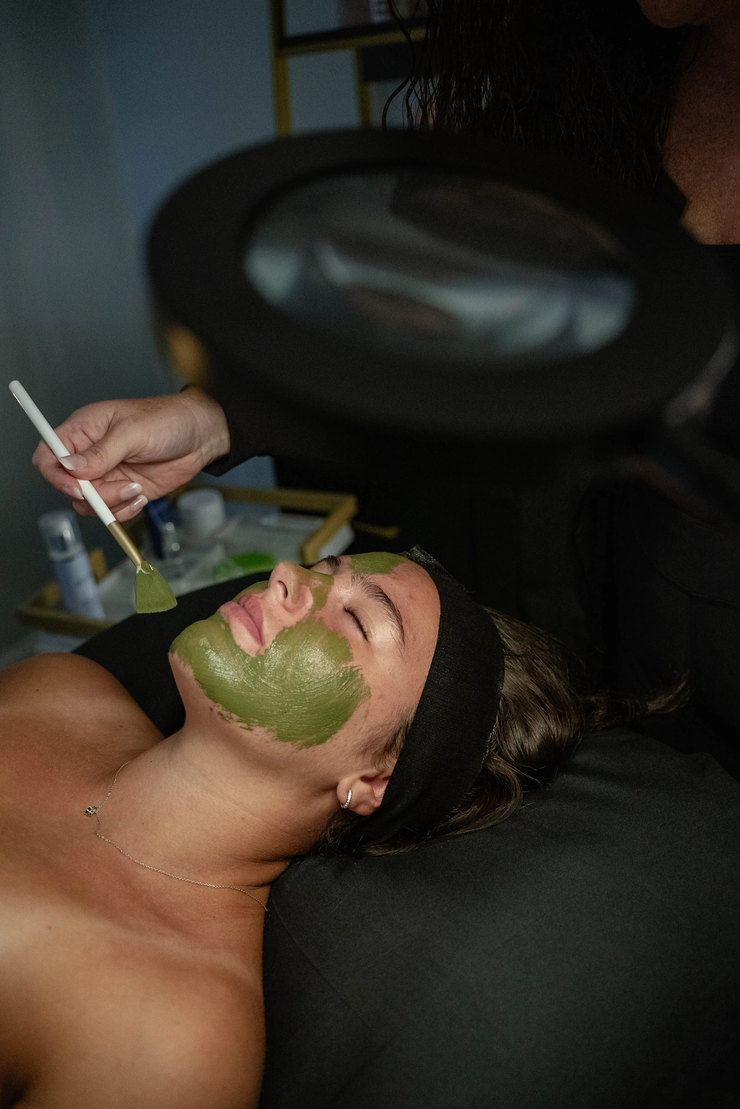 A woman lying on a treatment bed with her eyes closed, getting a facial treatment with green mask being applied by a practitioner using a brush. The woman is wearing a black headband and a necklace.