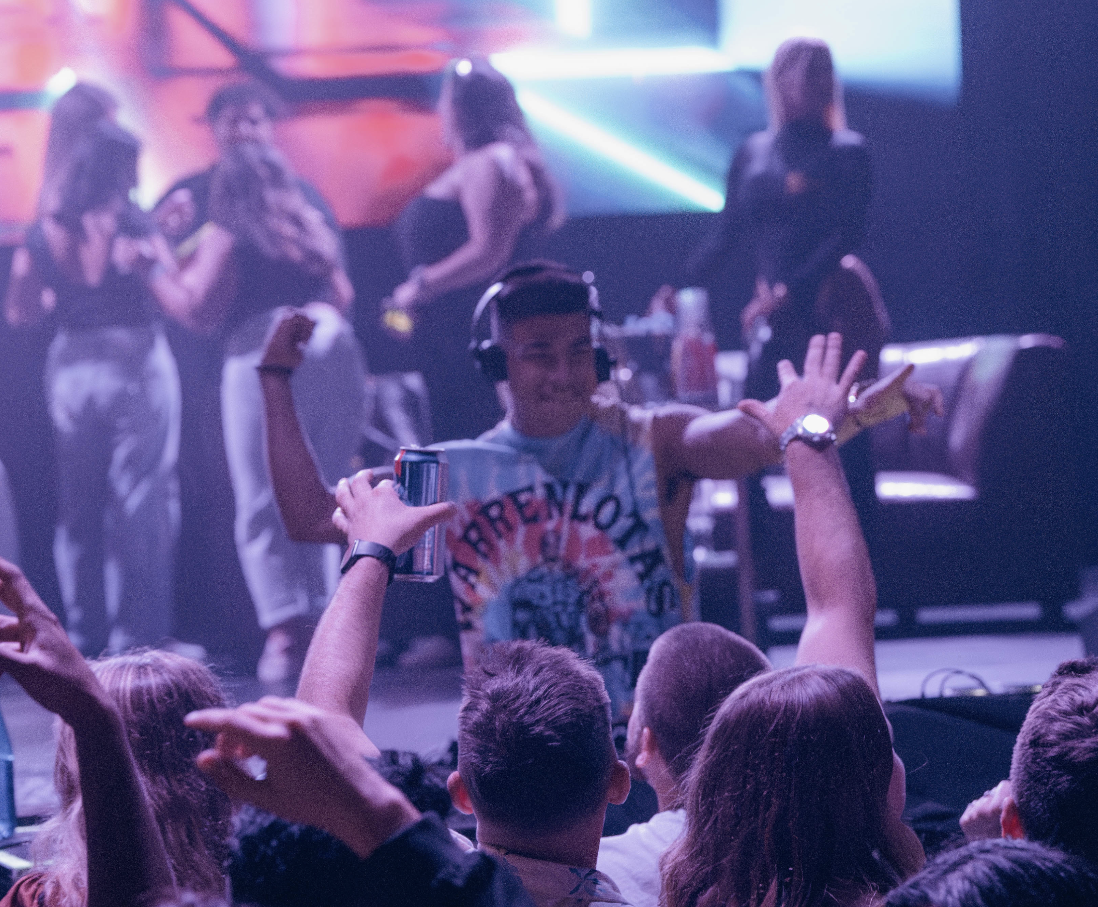 A person at a concert or event is dancing and smiling while wearing headphones and a colorful sleeveless shirt. The crowd around him is raising hands, and there are others in the background near a stage with bright, colorful lights.