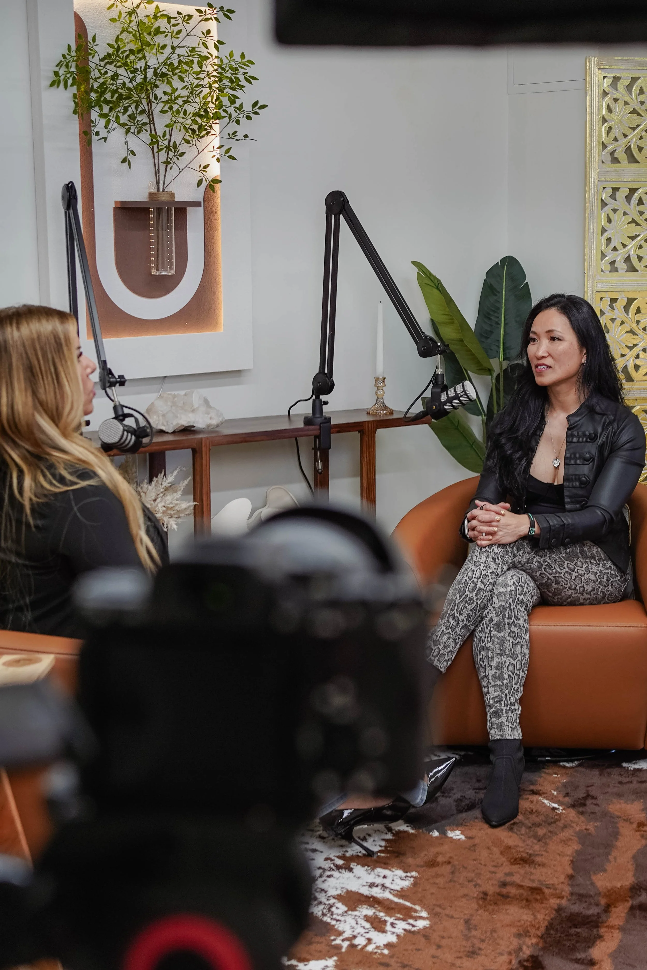 Two women having a conversation in a podcast studio with microphones, a camera in the foreground, and a decorative wall with plants in the background.
