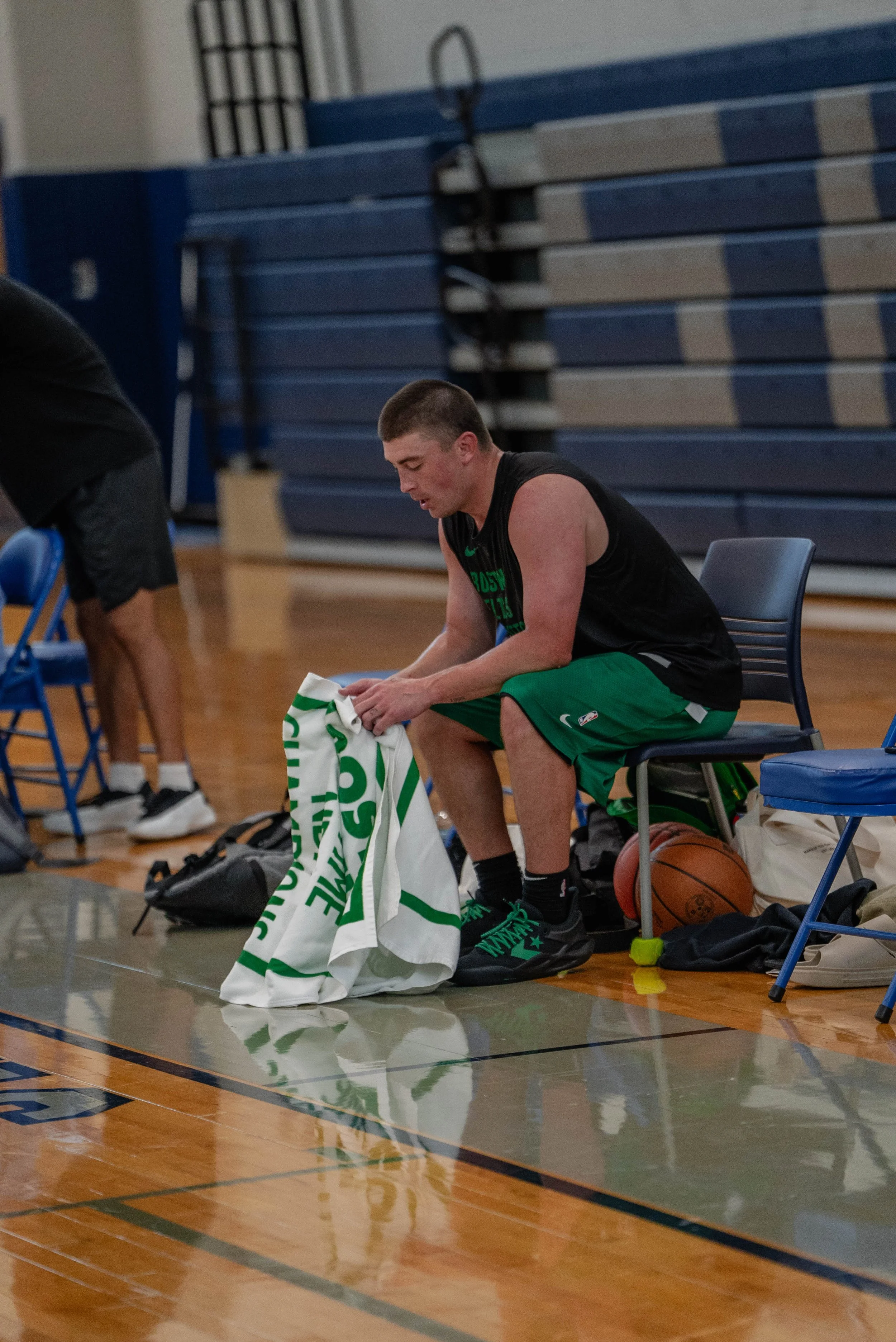 A young male basketball player sitting on a chair on a basketball court, putting on a towel. He is wearing a black sleeveless shirt and green shorts, with a basketball beside him. In the background, there are blue bleachers and another person leaning over a chair.
