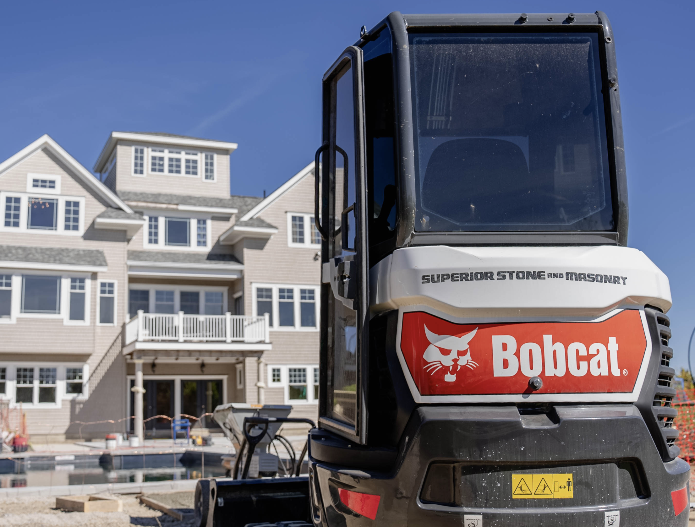 Construction site with a Bobcat skid-steer loader in the foreground and a large multi-story residential house under construction in the background under a clear blue sky.