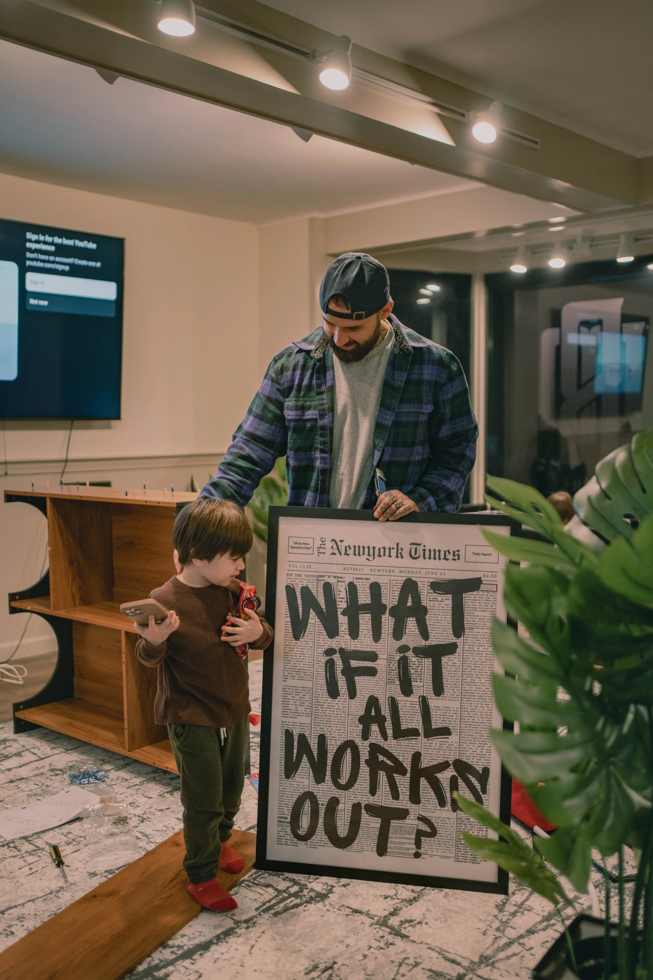 A man and a young boy standing near a framed newspaper with a question written on it, inside a room with a large flat-screen TV and a bookshelf.
