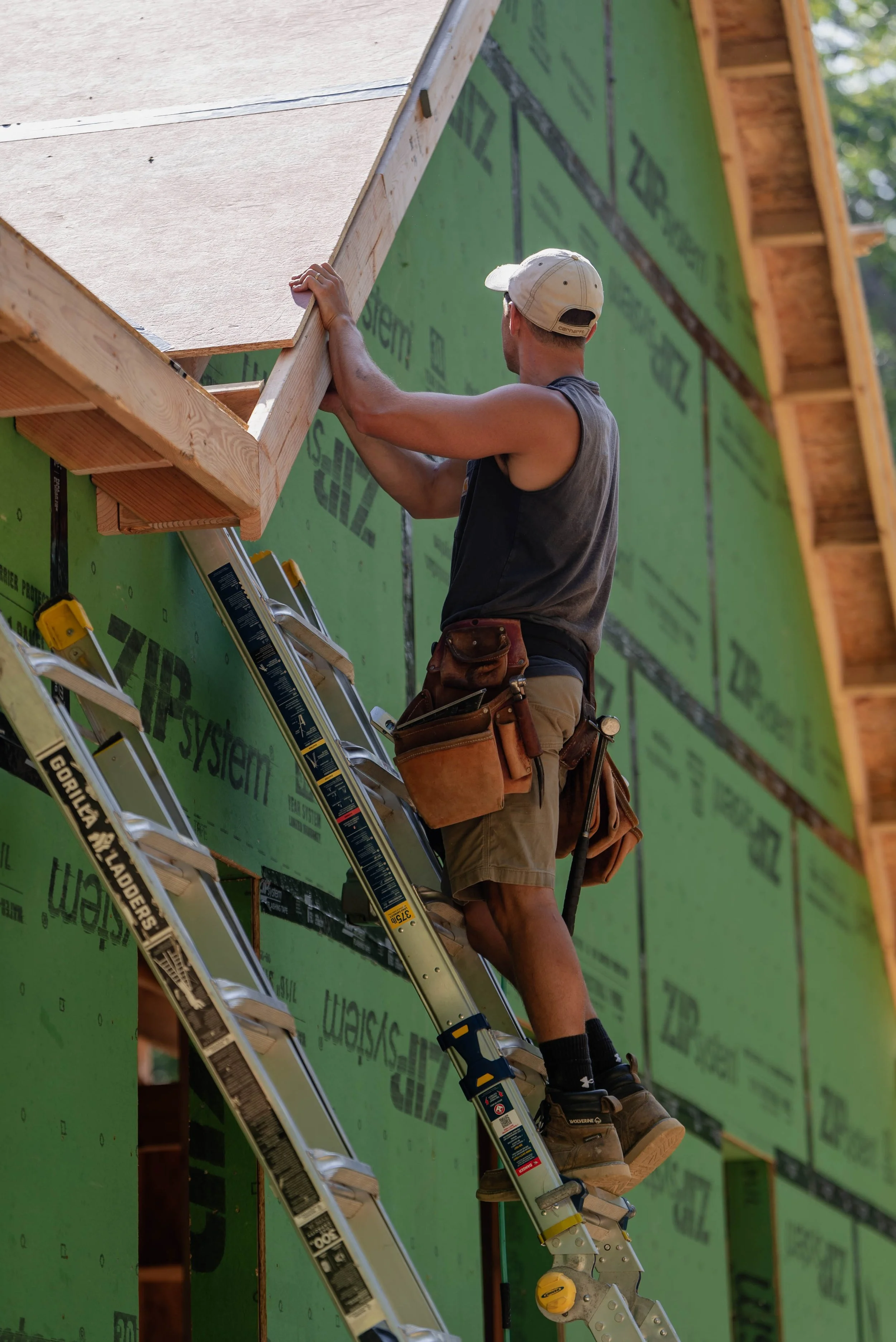 Construction worker installing a roof on a building, standing on a ladder with tools around his waist.