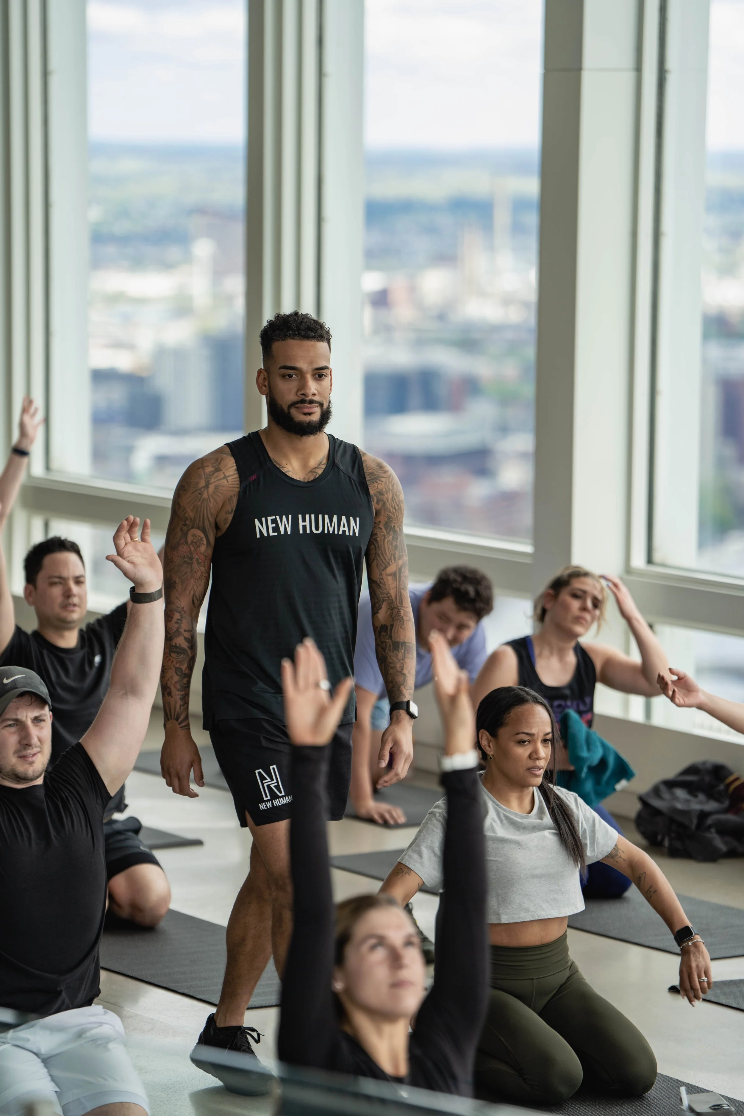 A diverse group of people participating in a yoga or stretch class in a high-rise studio with large windows, seated or kneeling on mats, some raising hands or touching their heads, while an instructor stands observing.