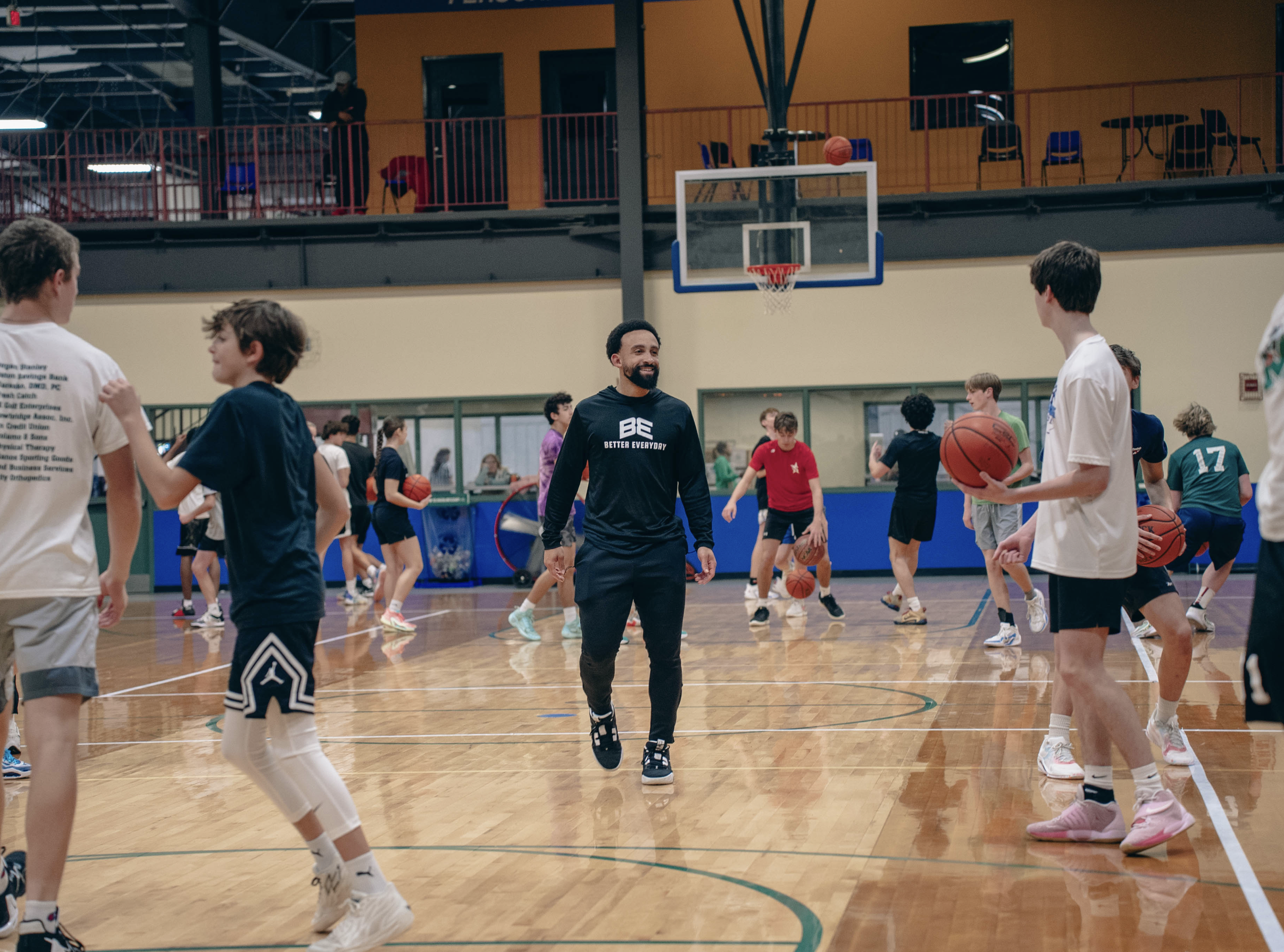 People playing basketball in an indoor gym, with a coach or instructor in the center smiling and watching kids practice dribbling and shooting basketballs.