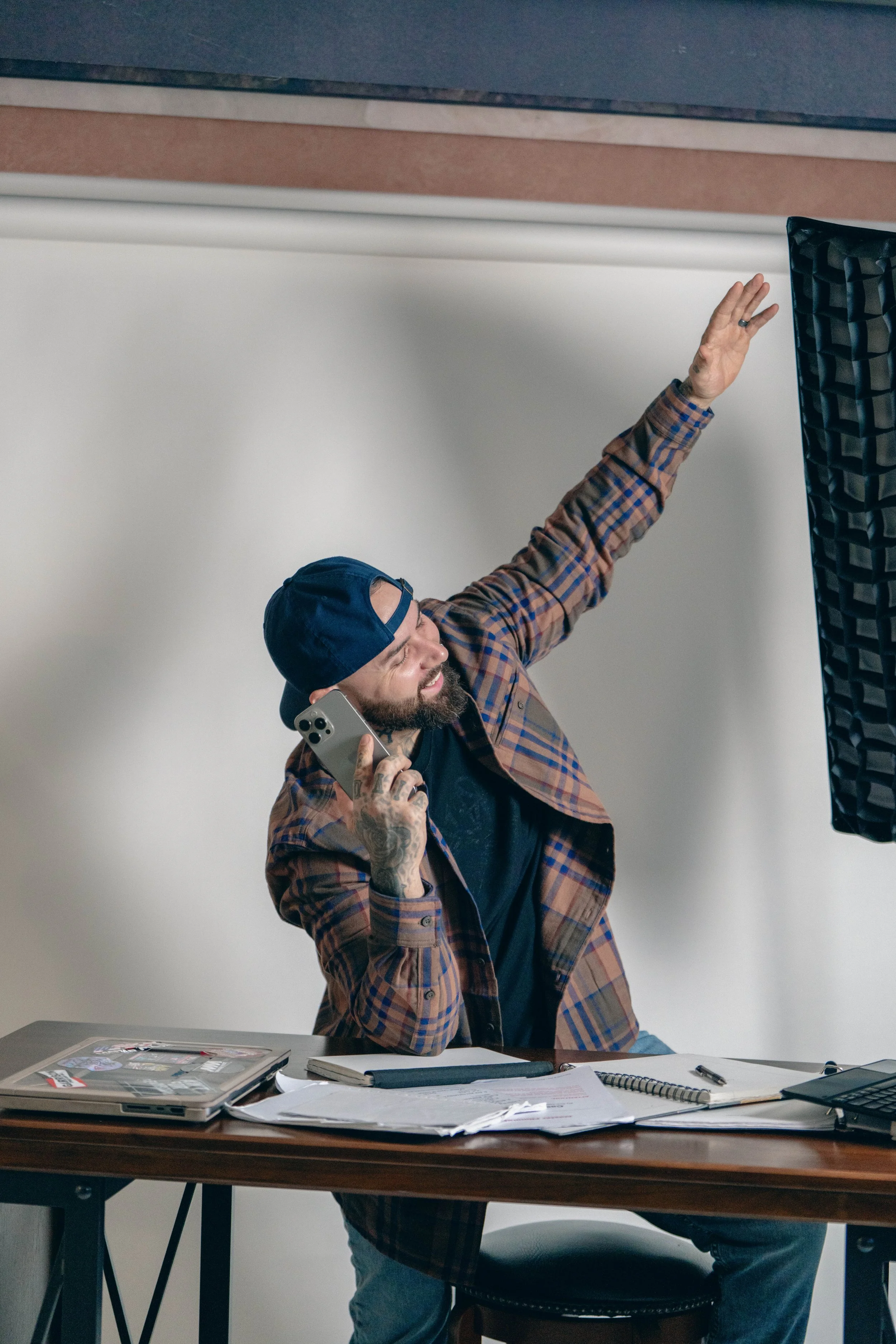 A man with a beard, tattooed hand, wearing a backwards baseball cap and plaid shirt, sitting at a desk, talking on the phone, smiling, and raising his left hand.