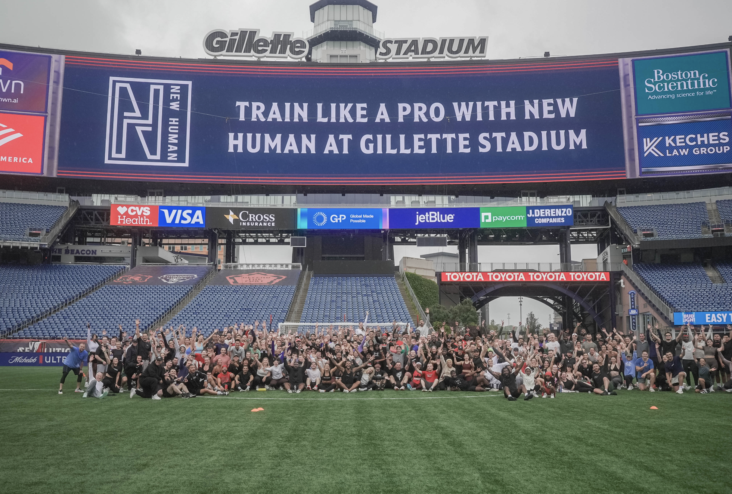 A large group of people on the field of Gillette Stadium, celebrating and posing for a photo, with a big electronic scoreboard overhead displaying an advertisement and the message, "Train like a pro with new human at Gillette Stadium." The stadium has blue seats and advertisements along the upper levels.