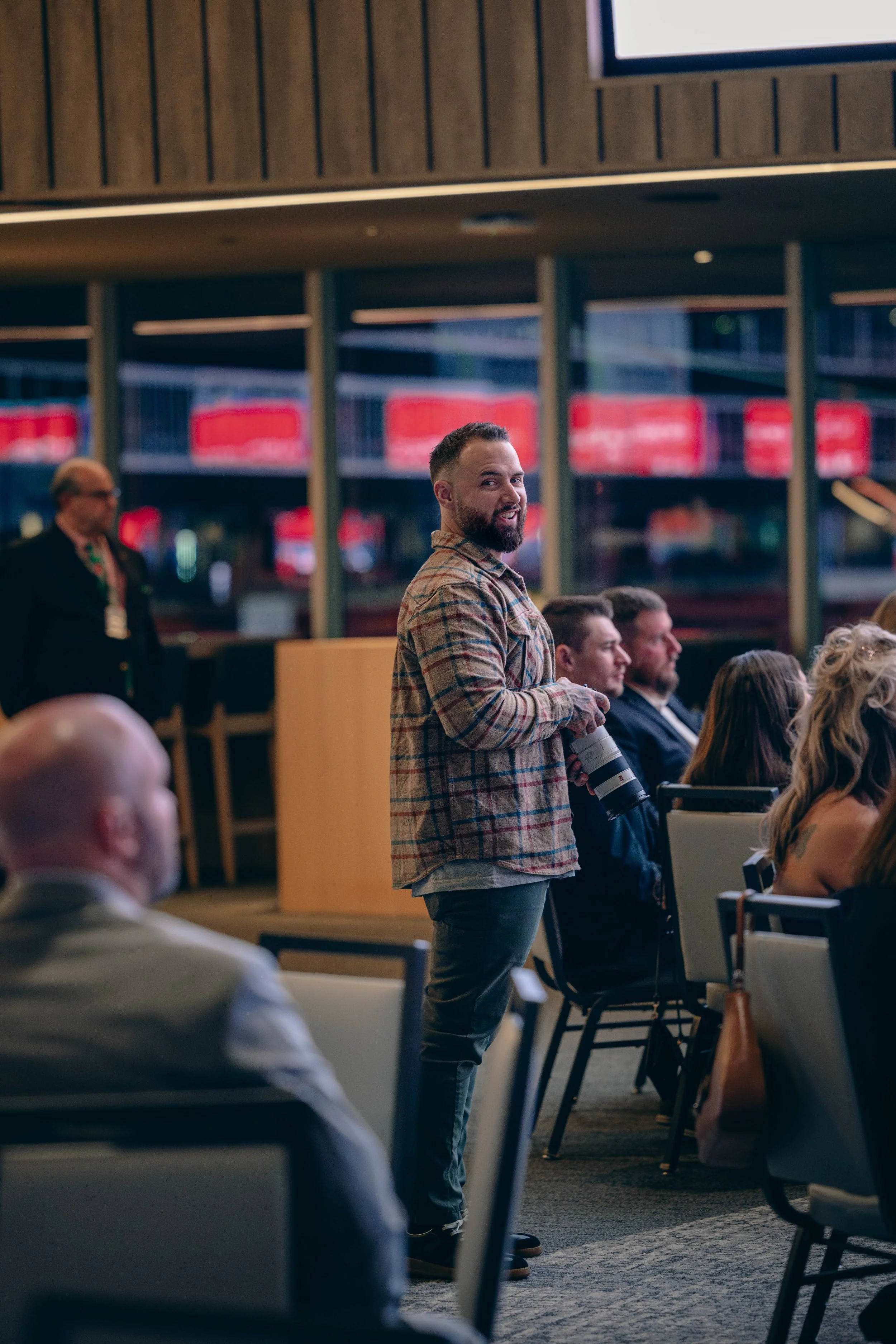 A man with a beard, wearing a plaid shirt, holding a camera, standing and looking towards the camera, in a conference or meeting room with other seated attendees, some men in suits, and a woman with curly blonde hair, a woman with a tattoo, and a man with a gray beard, in front of large windows.