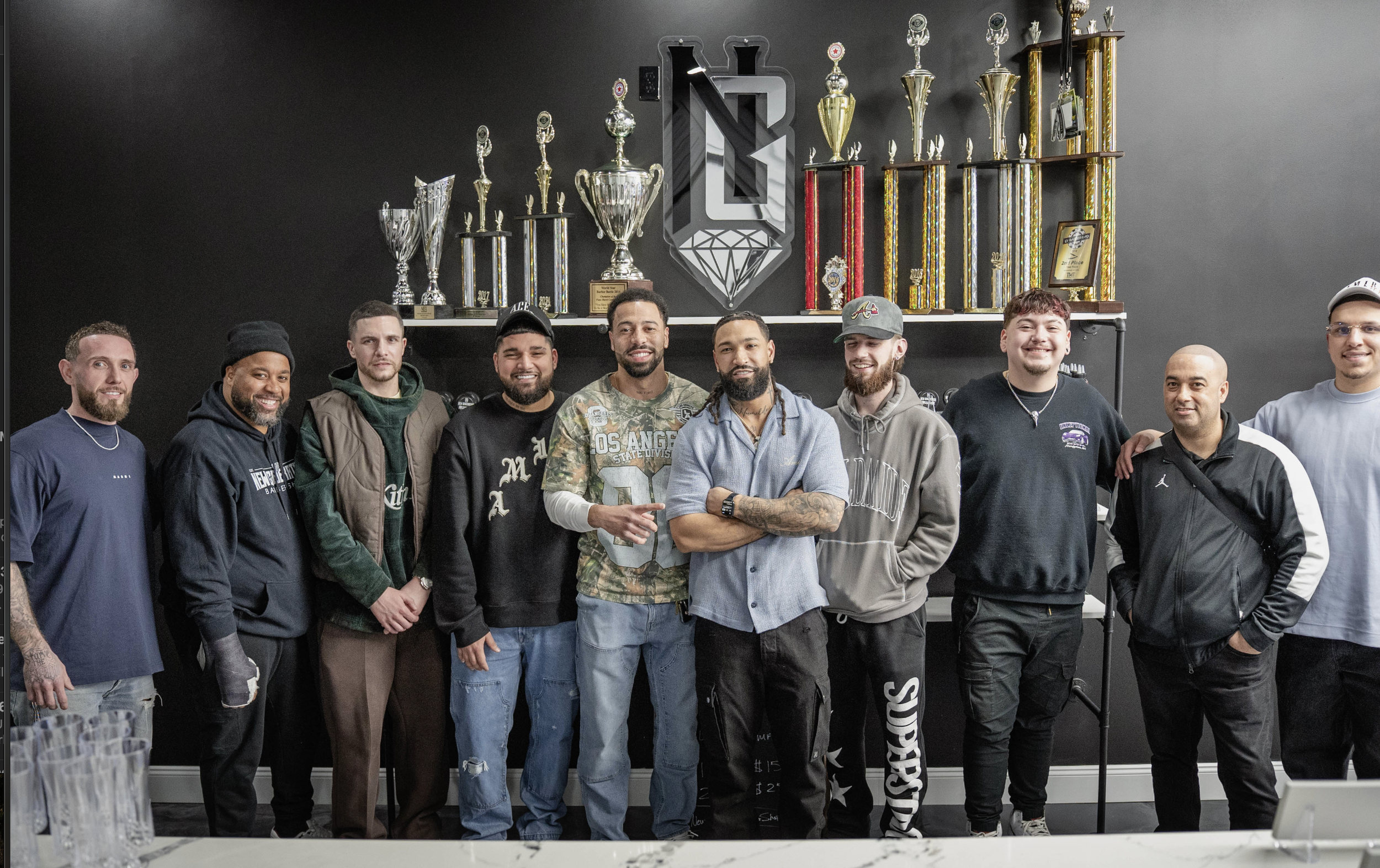 Group of ten men standing in front of a wall decorated with trophies and a logo, posing for a photo.