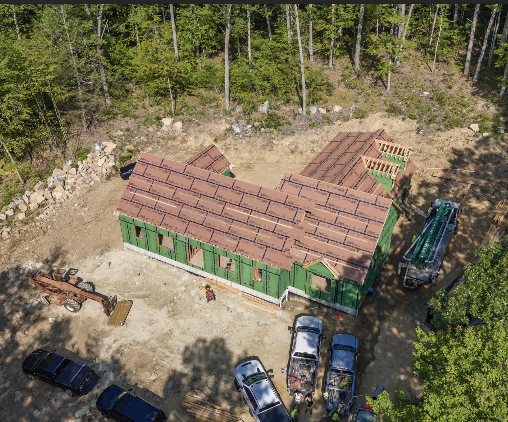Aerial view of a house under construction with green sheathing and a brown roof, surrounded by construction vehicles and workers, with a wooded area in the background.