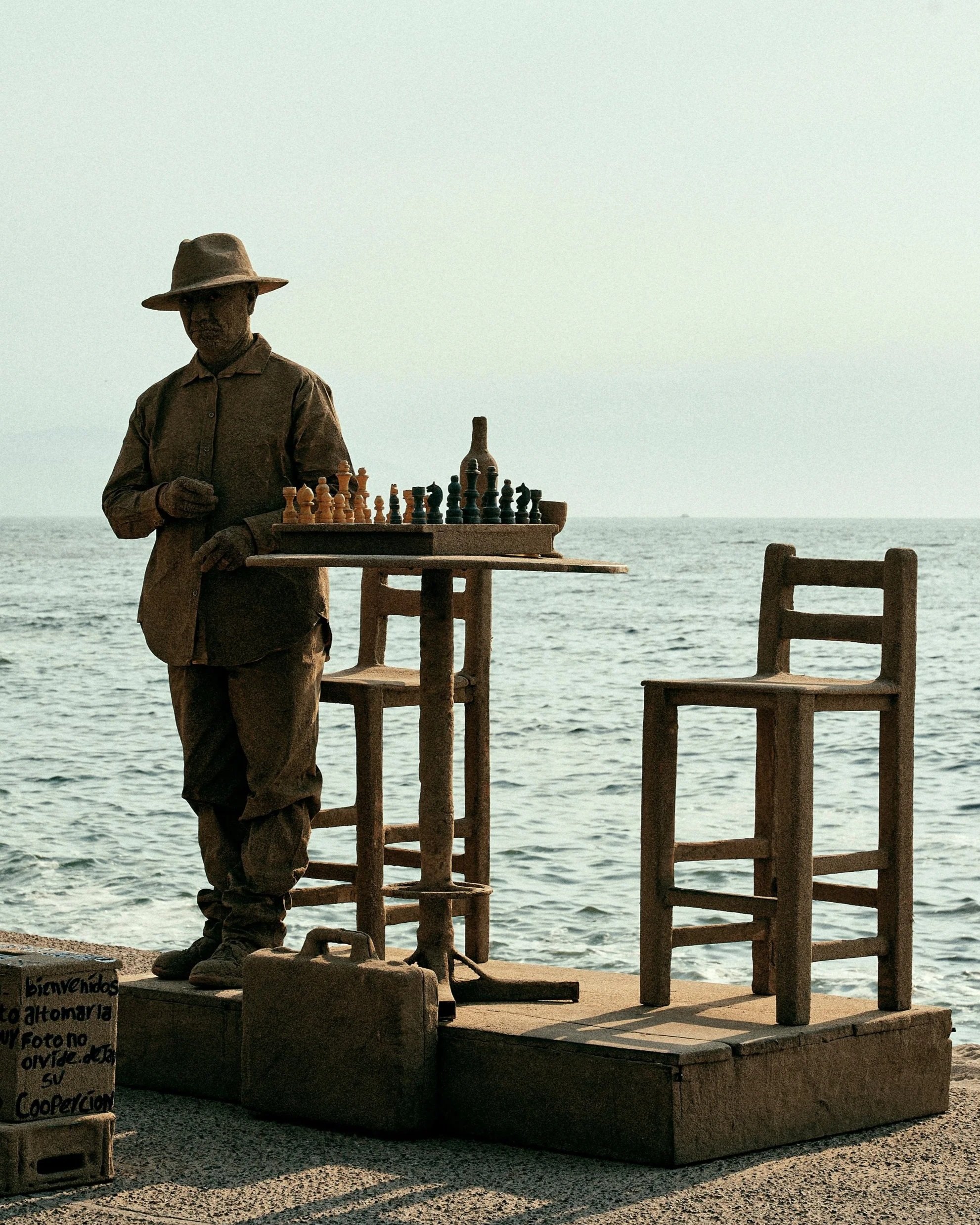 A statue of a man playing chess outdoors by the sea, with two wooden chairs nearby and a sign in Spanish on the ground.