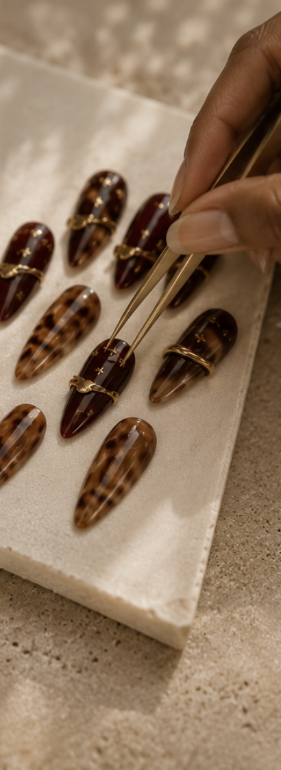 Person using tweezers to place jewelry on a marble surface with tortoiseshell nail charms and gold rings.