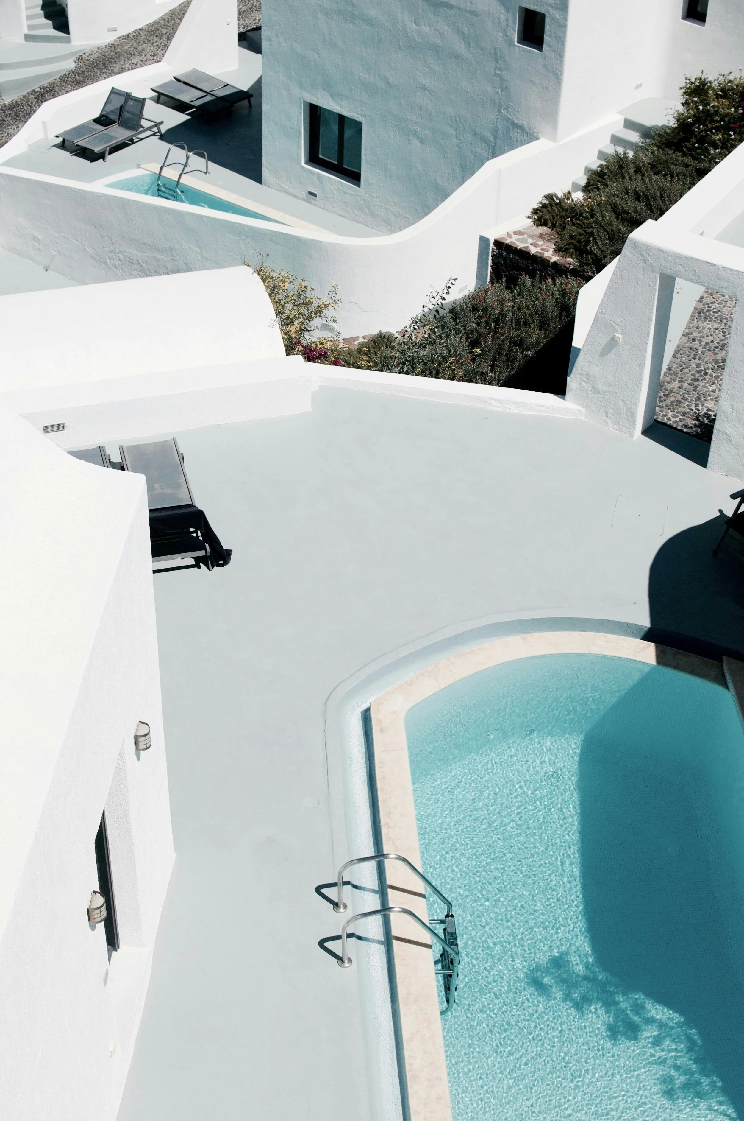 Aerial view of a white building complex with a small swimming pool, lounge chairs, and steps leading to other levels, in a coastal Mediterranean style.
