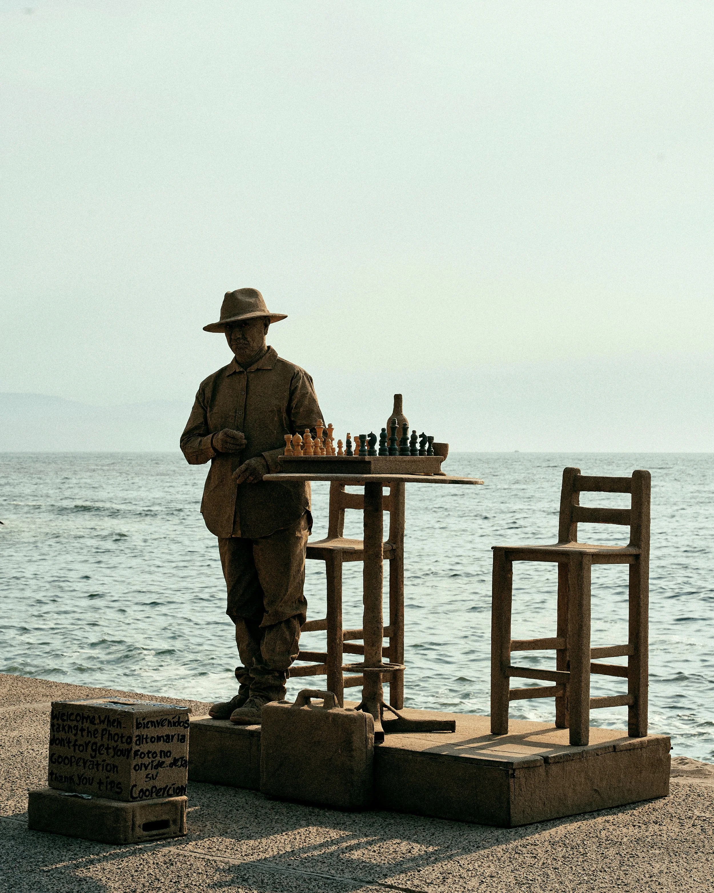 A street performer dressed as a scarecrow playing chess by the sea with empty chairs and a sign with messages in multiple languages.